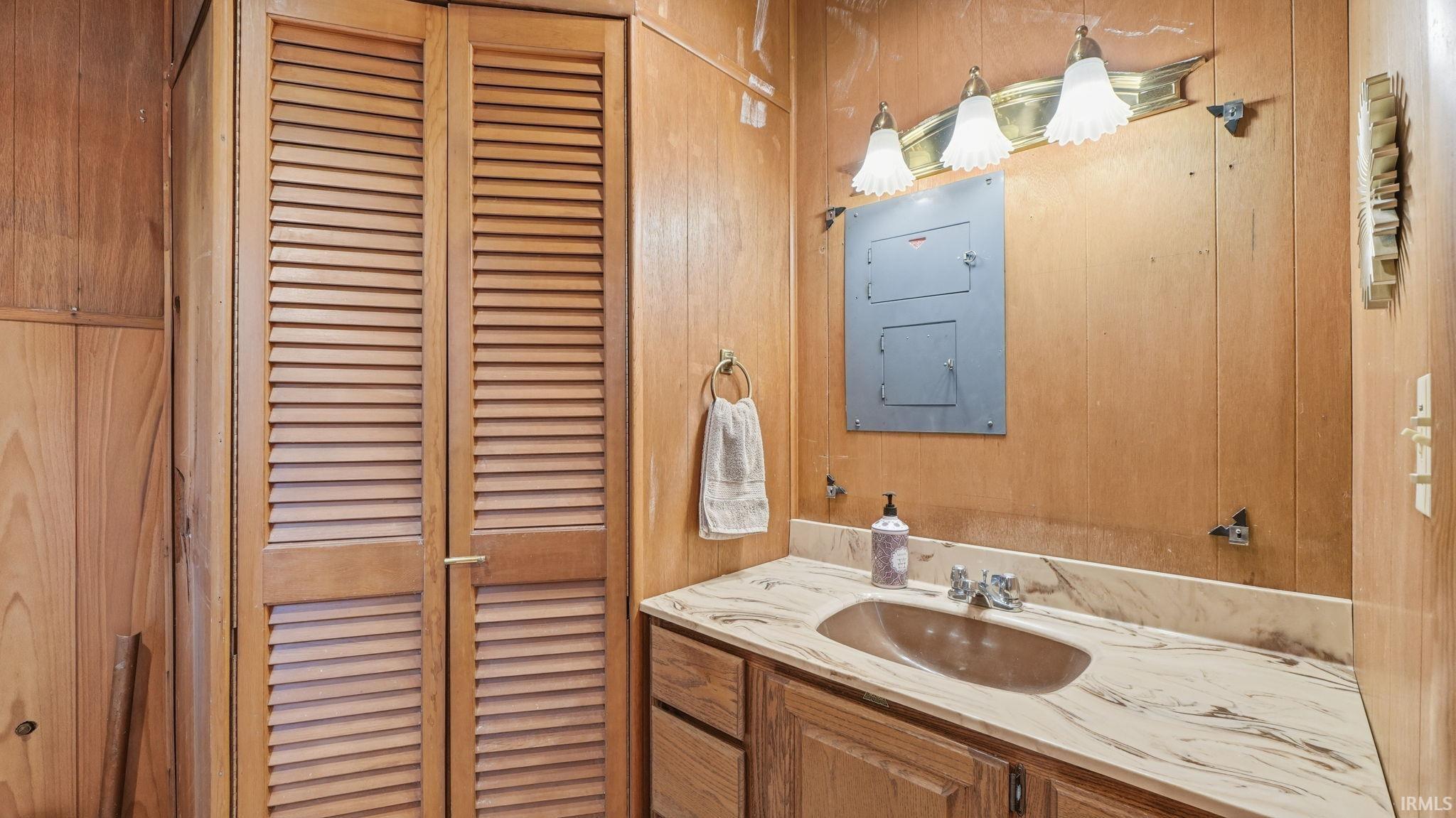 Wood-paneled room featuring louvered bi-fold doors, a vanity with a patterned countertop and integrated sink, a dual-handle faucet, and an overhead three-bulb light fixture