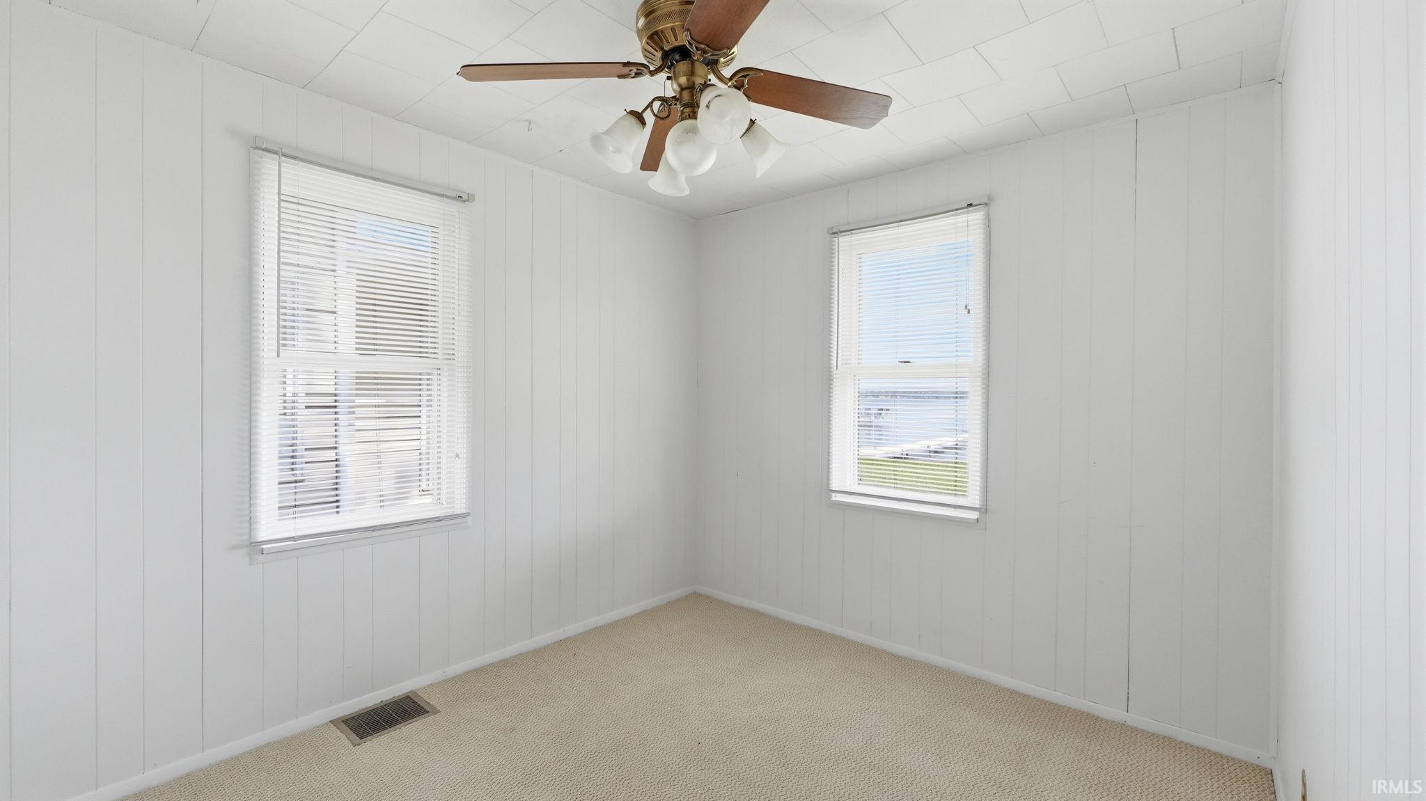 Bright room featuring two windows with horizontal blinds, a ceiling fan with light fixture, white vertical paneling, and neutral carpeting