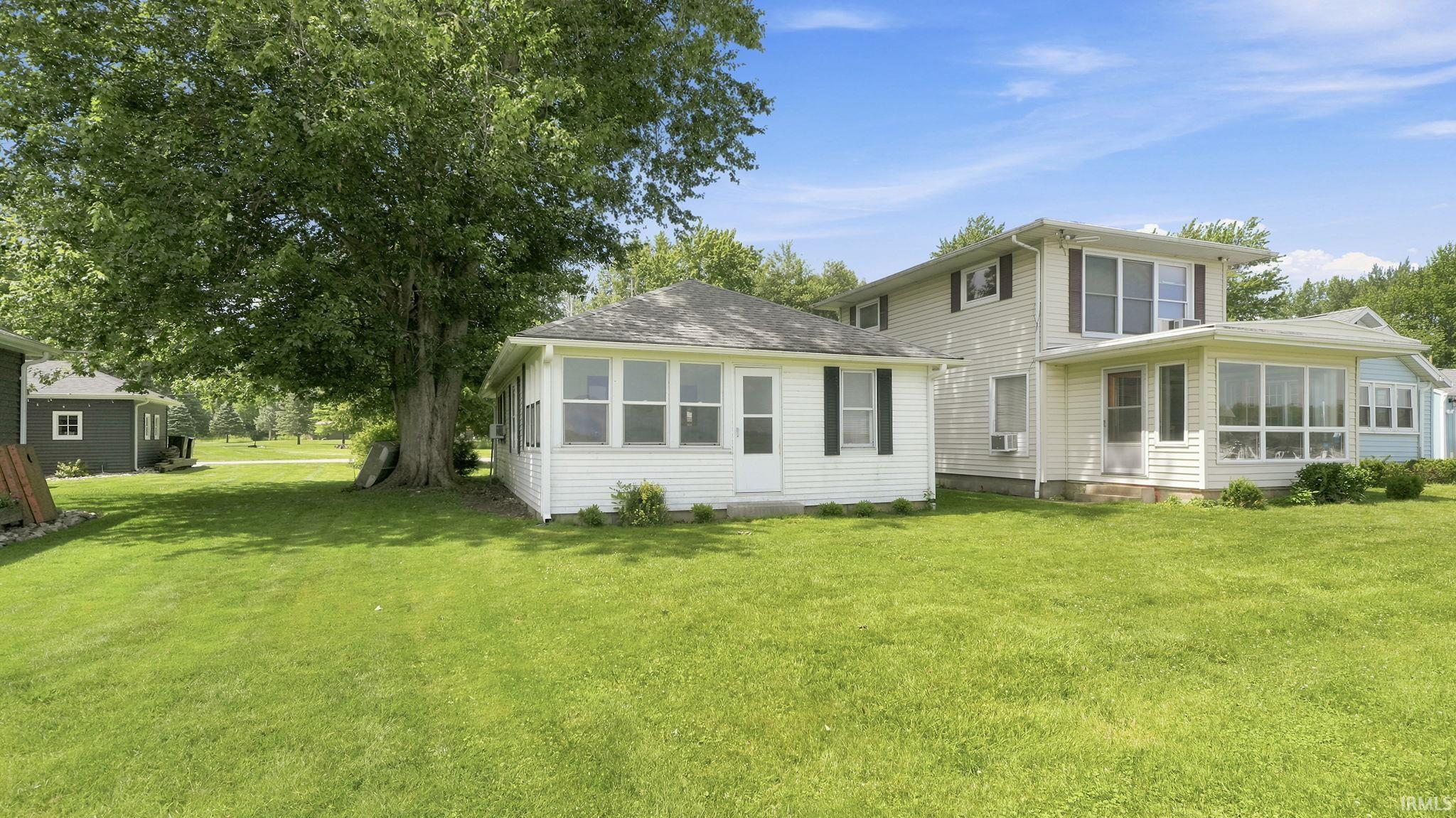 Expansive green lawn with mature shade trees, showcasing a multi-level white siding residence featuring a sunroom addition and a detached white outbuilding