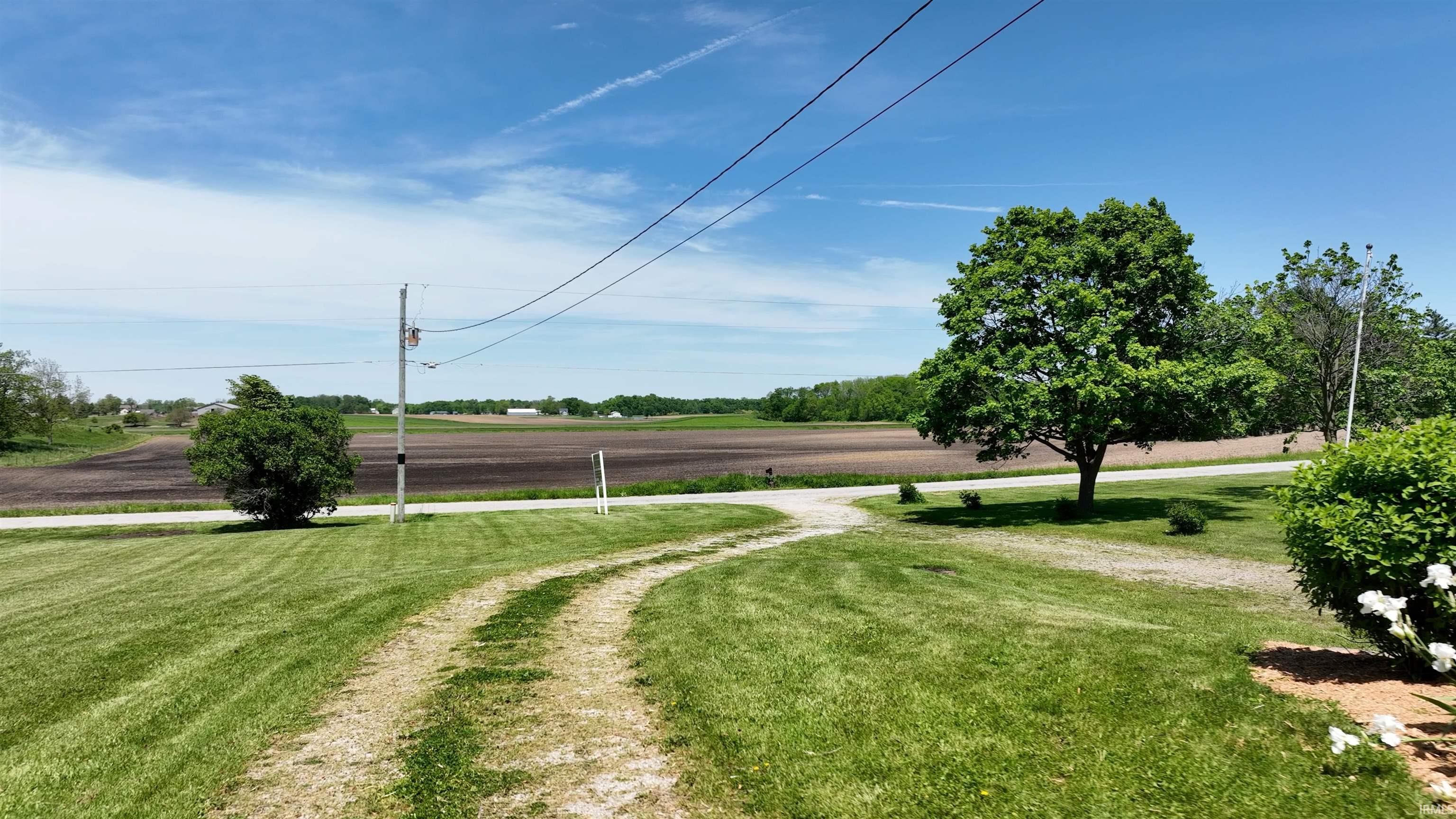 Extensive green lawn with a curving gravel driveway, mature trees, and distant cultivated fields under a clear blue sky