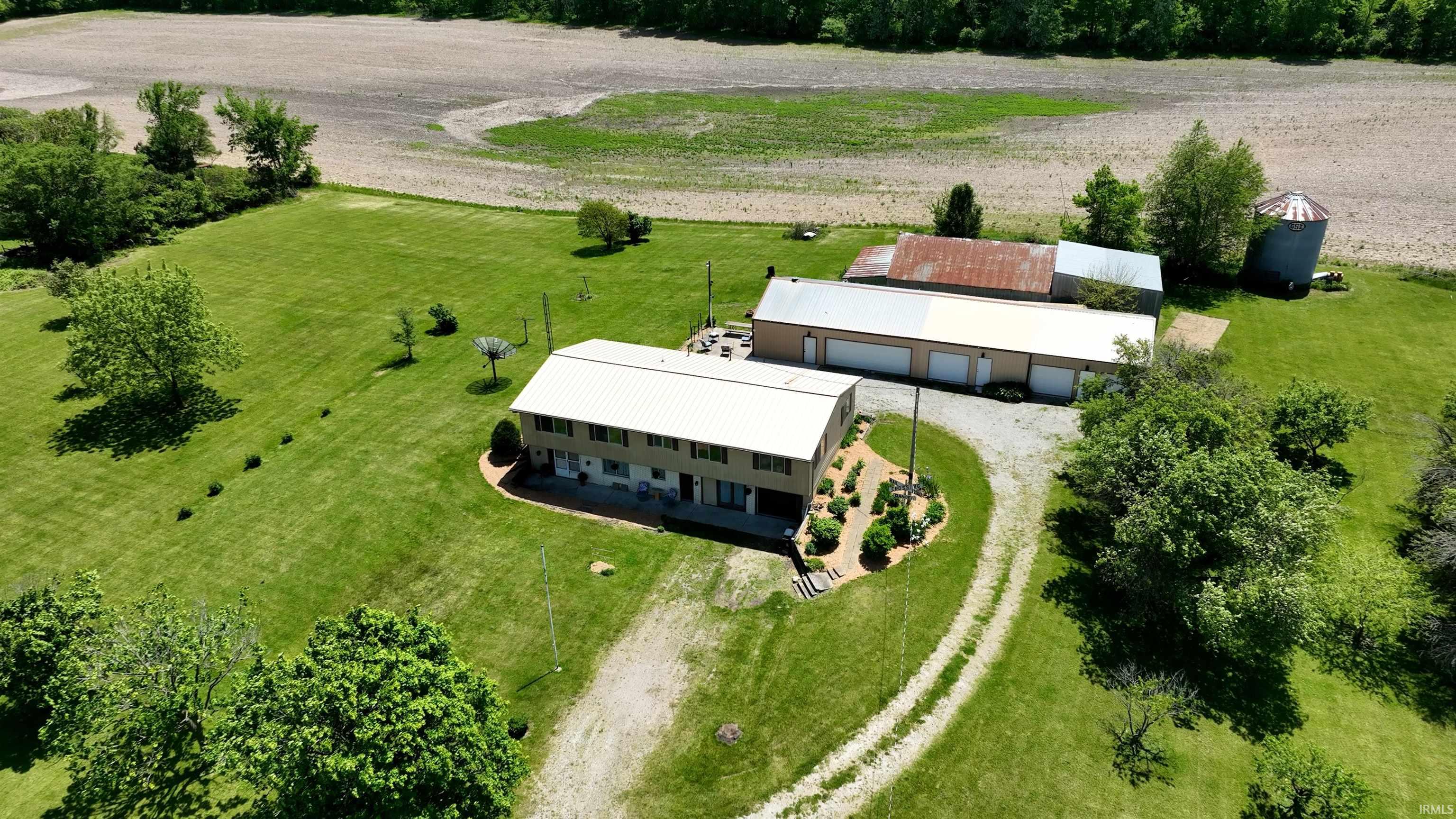 Sprawling property featuring a multi-level residence with a metal roof, extensive gravel driveway, large detached garage structure with multiple bay doors, mature trees, and a silo