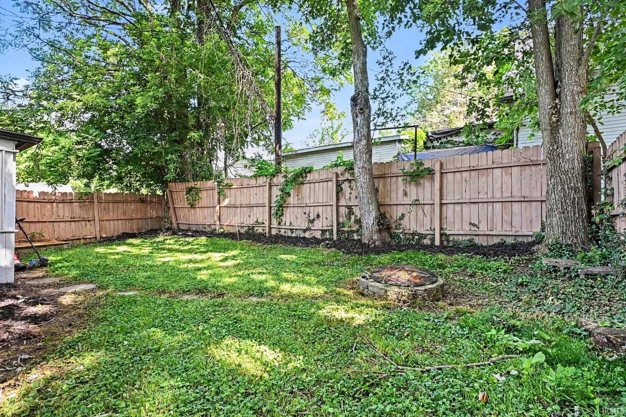 Private backyard featuring a wood privacy fence, mature trees, an established fire pit, and a grassy lawn area