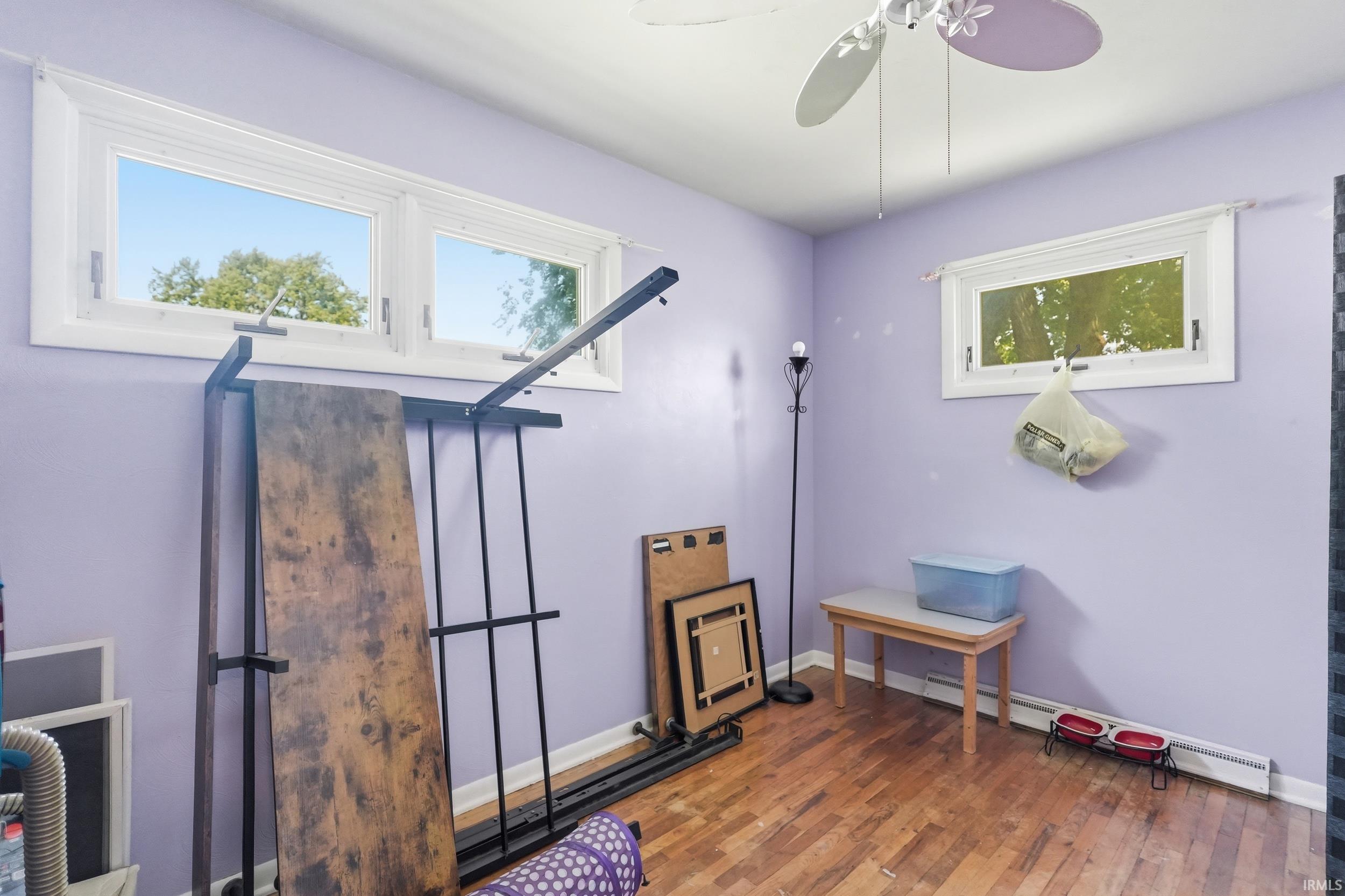 Room featuring wood-finish flooring, two large windows with white trim, a ceiling fan with lavender blades, and lavender-painted walls
