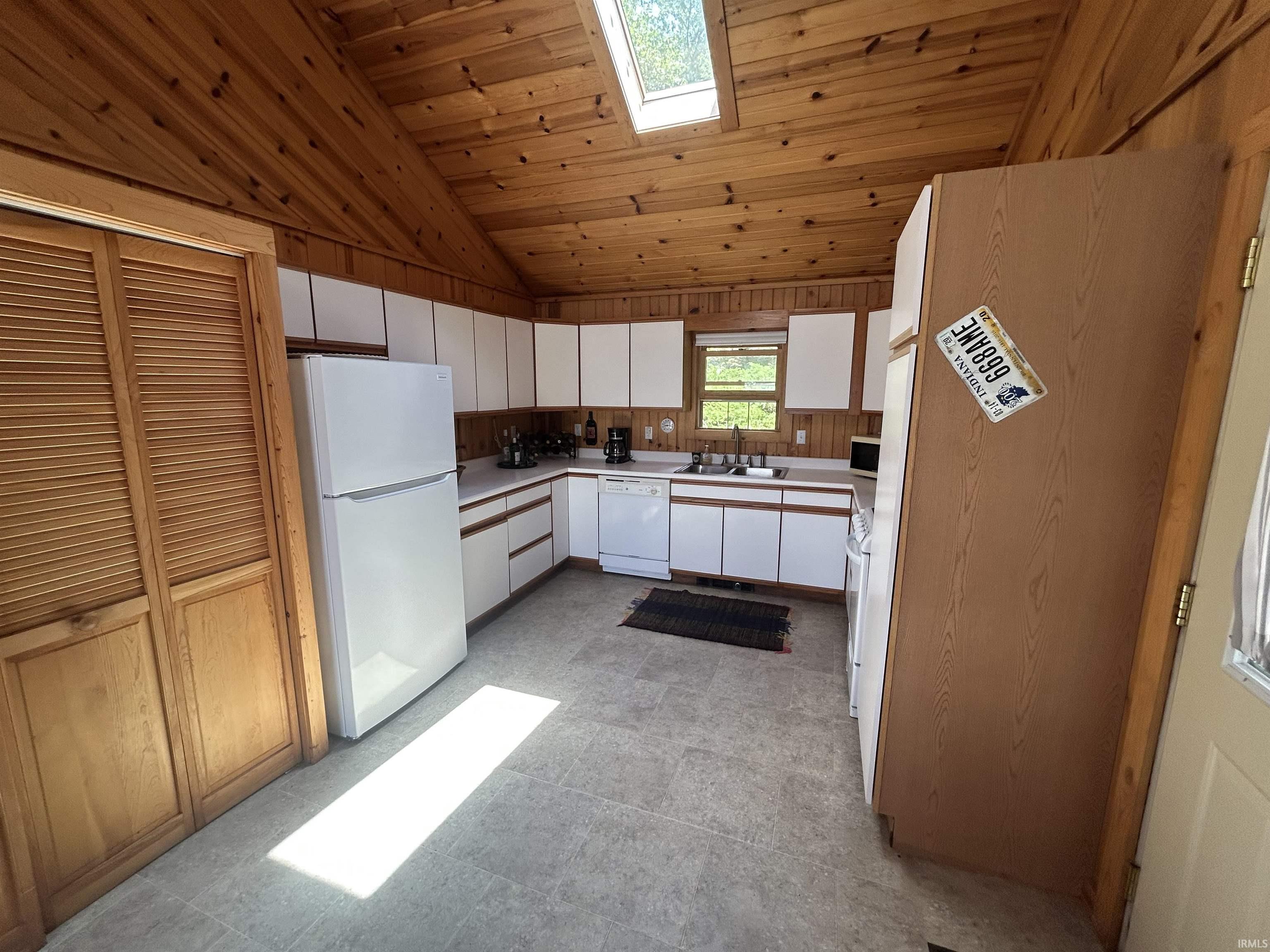 Kitchen featuring wood-paneled vaulted ceiling, white cabinetry with wood-finish trim, a skylight, a window overlooking exterior greenery, and light-toned tile flooring