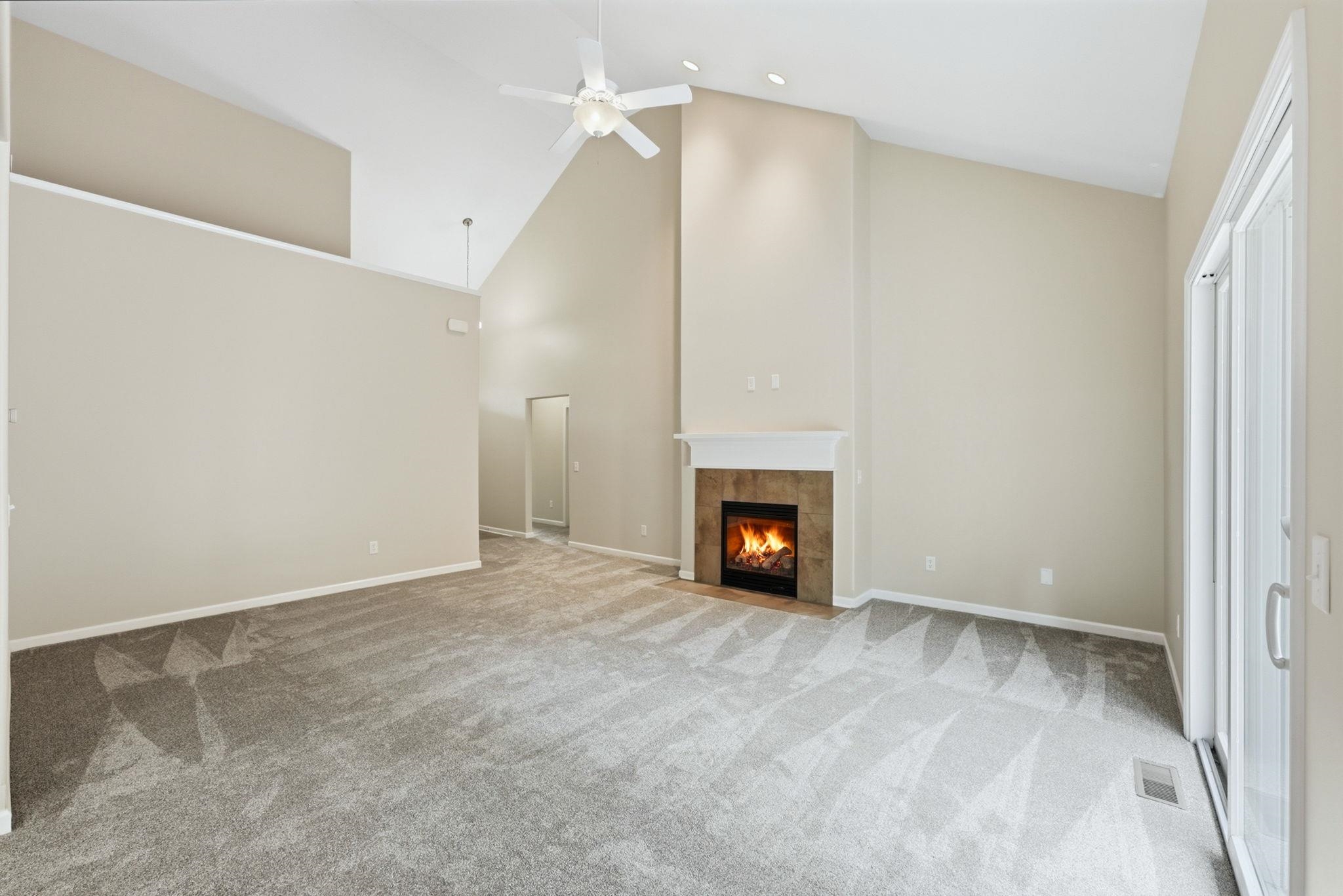 Vaulted ceilings featuring a ceiling fan, a fireplace with a mantel and tile surround, and light-toned carpeting throughout