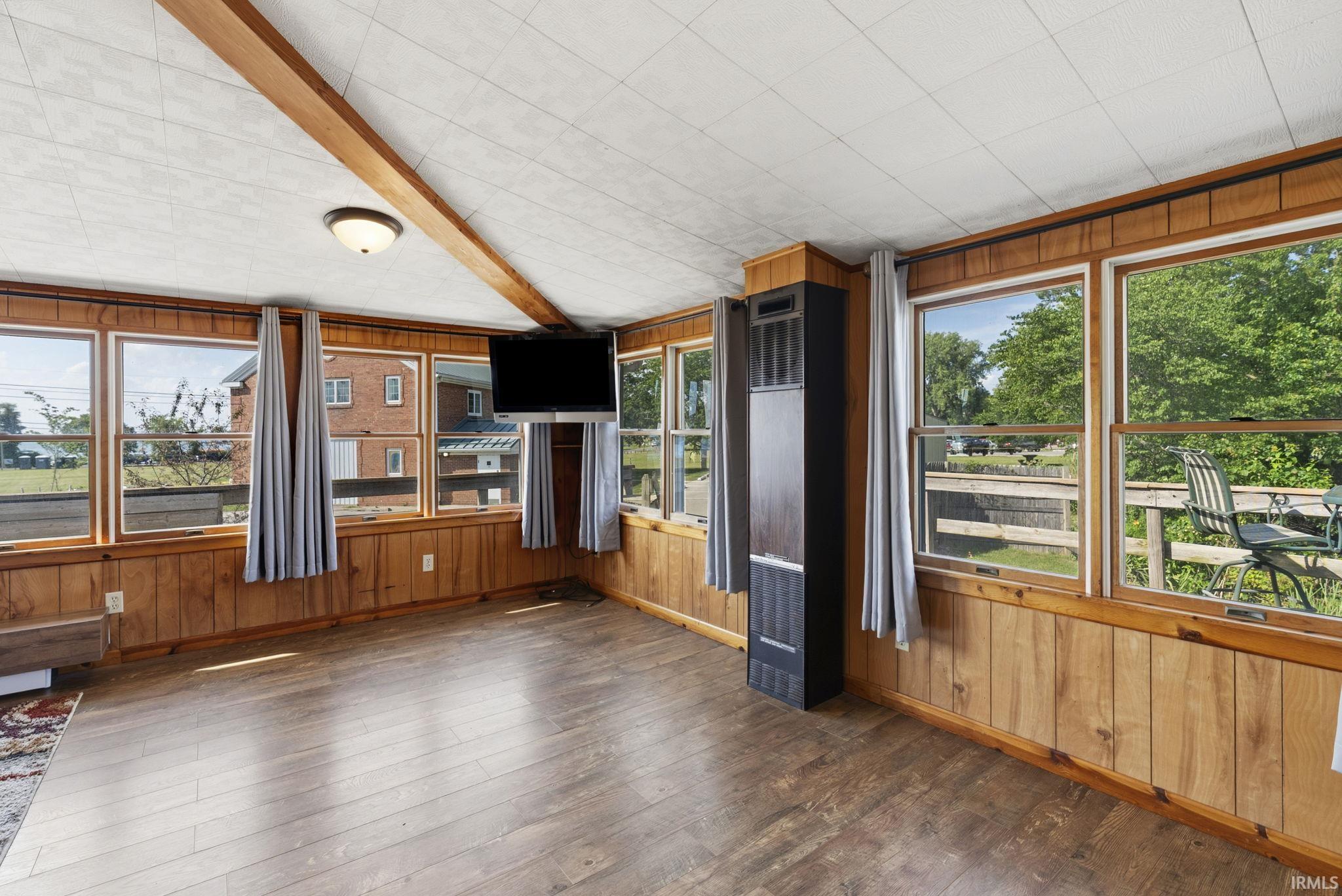 Sunroom featuring wood-finish flooring, wood paneling, multiple windows, exposed ceiling beam, and a flush mount light fixture