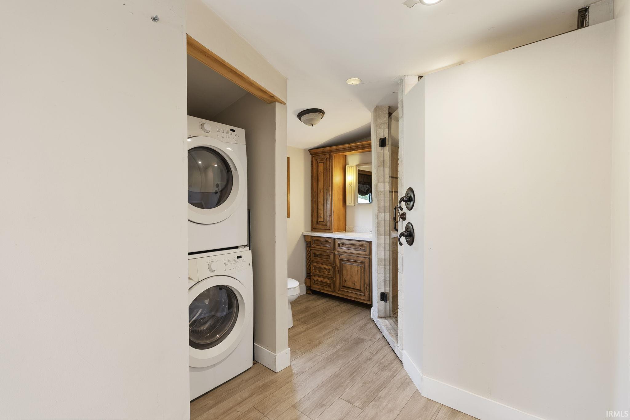 Compact laundry area featuring a stacked washer and dryer, wood-finish flooring, a vanity with wood cabinetry, and a shower with dark bronze fixtures