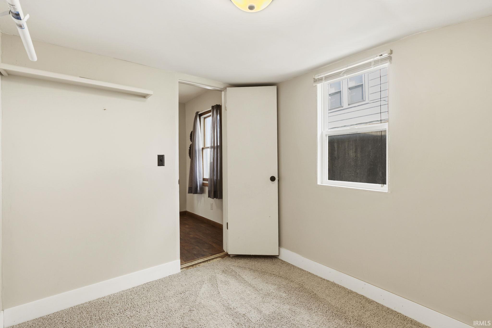 Carpeted room with a single pane window featuring white blinds, neutral wall paint, white trim, and a flush-mount ceiling light