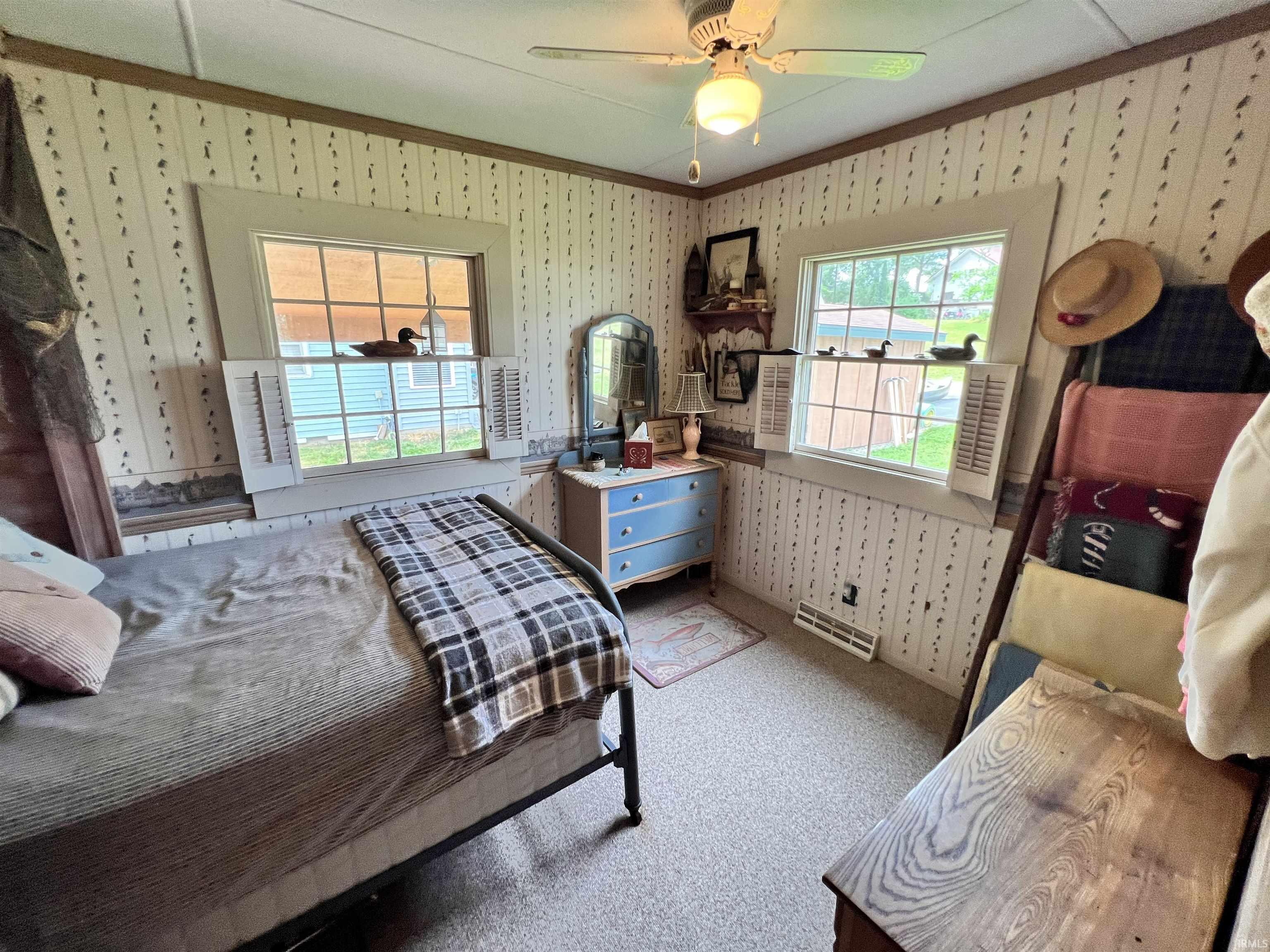 Room featuring two windows with grid detailing and interior shutters, textured wallpaper, carpet flooring, a ceiling fan with integrated lighting, and a metal bed frame