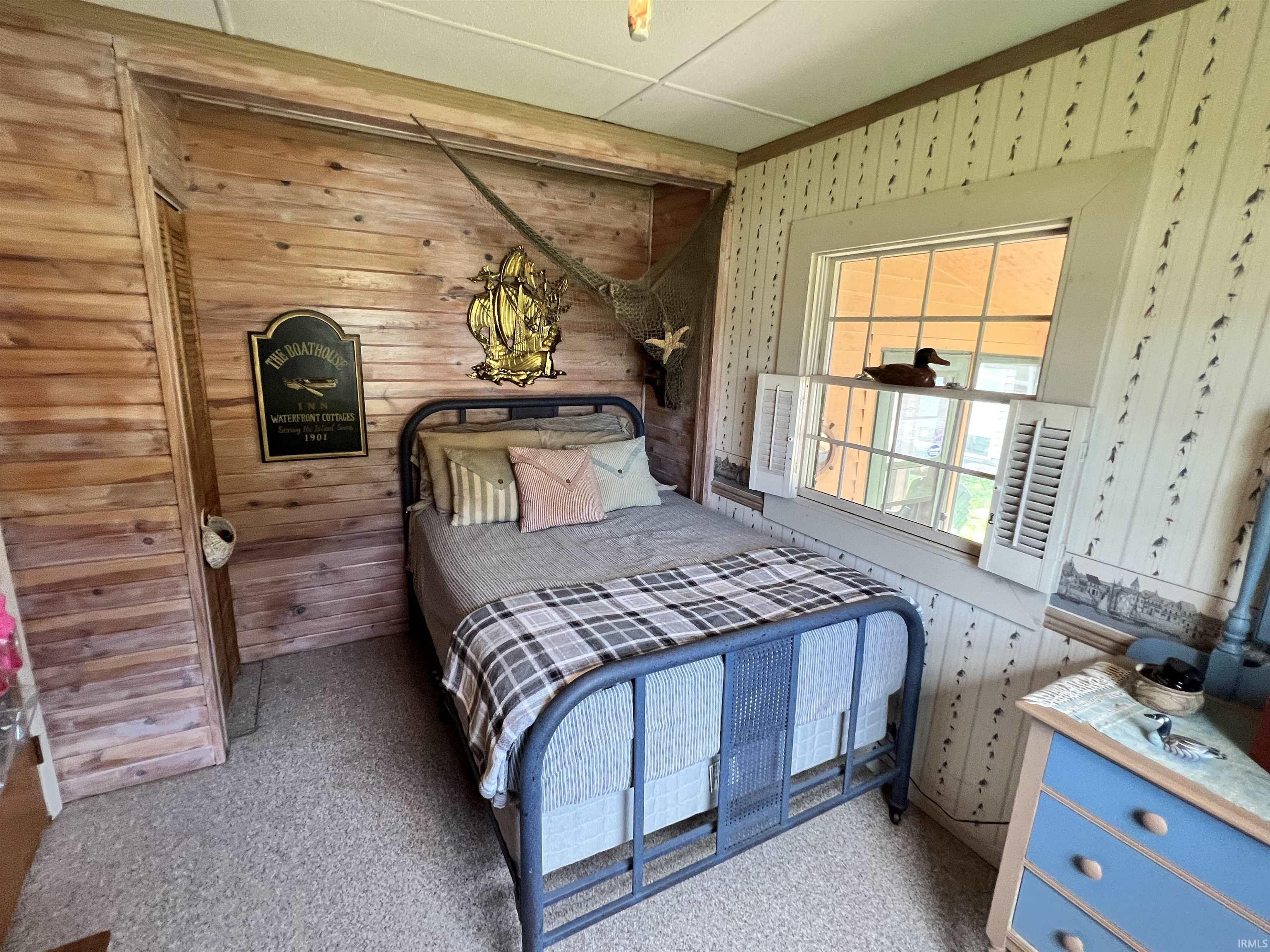Bedroom featuring carpet flooring, wood plank accent wall, built-in closet with louvered door, and a window with louvered shutters