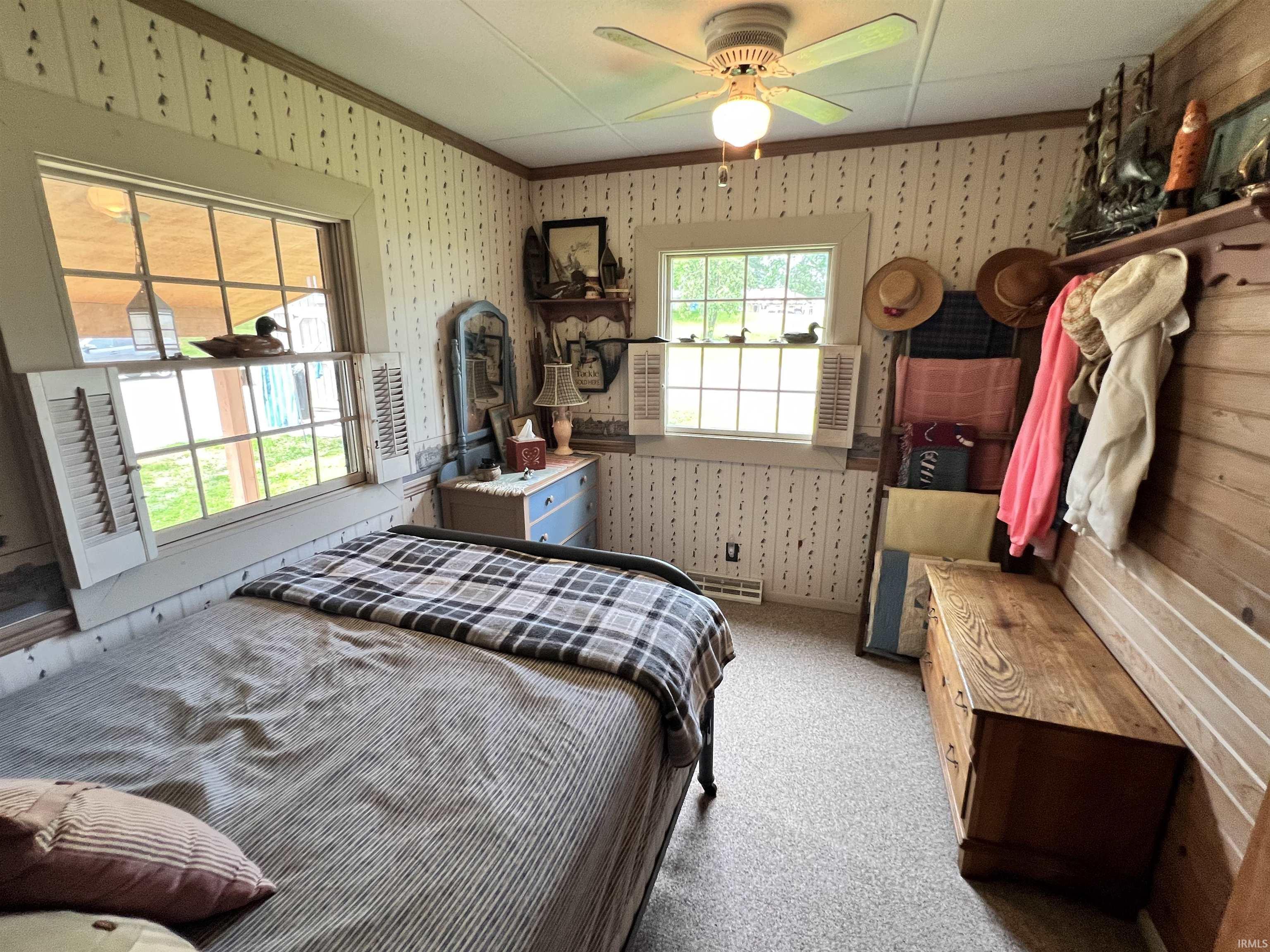 Bedroom featuring dual windows with white interior shutters, a ceiling fan with integrated lighting, wood-finish wall paneling, and patterned wallpaper