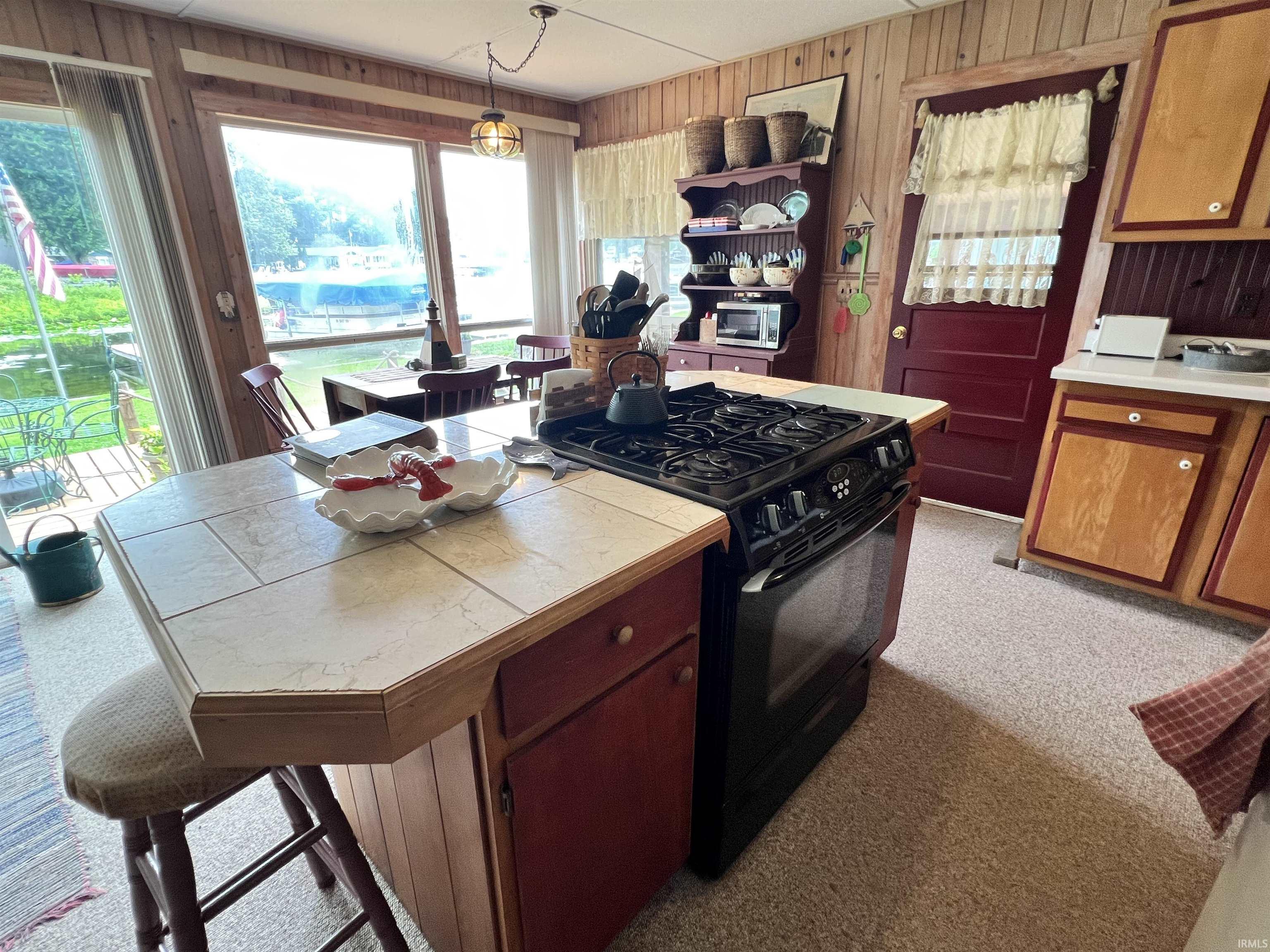 Kitchen featuring an island with tile countertop and integrated cooktop, wood paneling, wood cabinetry, and a full-light window overlooking a body of water