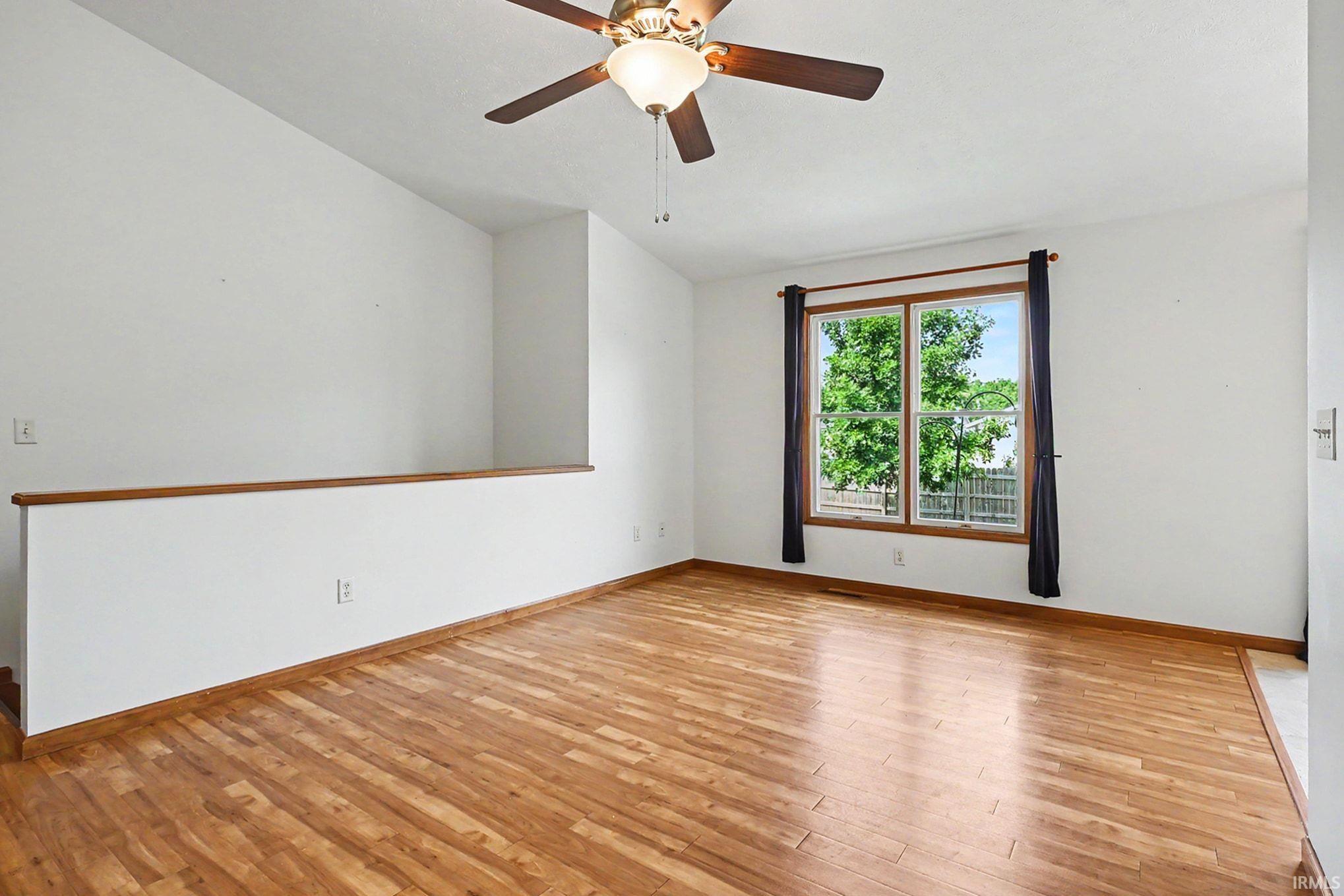 Spacious room featuring wood-finish flooring, a ceiling fan, and a large window with wood trim