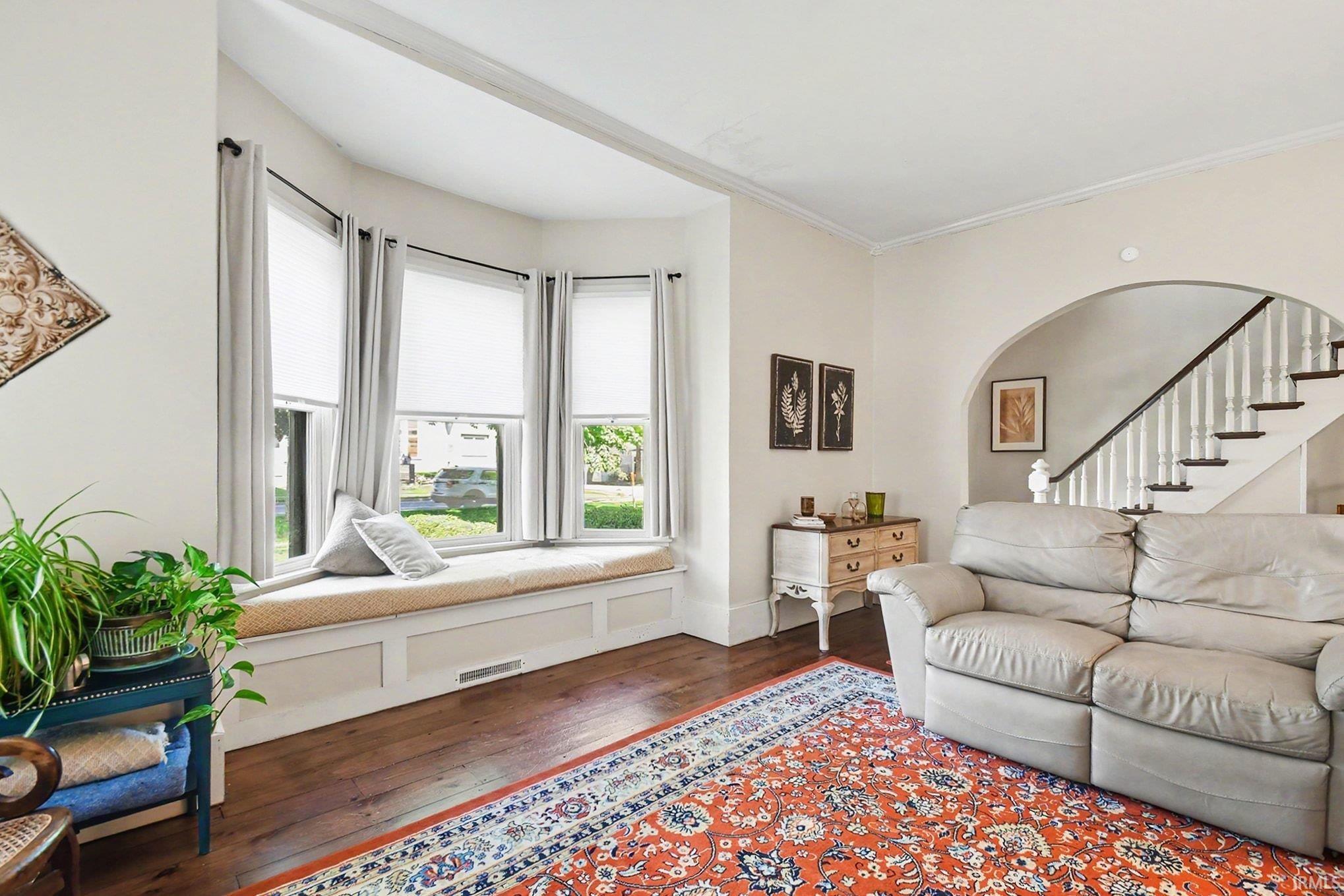 Bay window with integrated window seat, wood-finish flooring, white trim details, arched doorway, and staircase with white risers and dark wood handrail