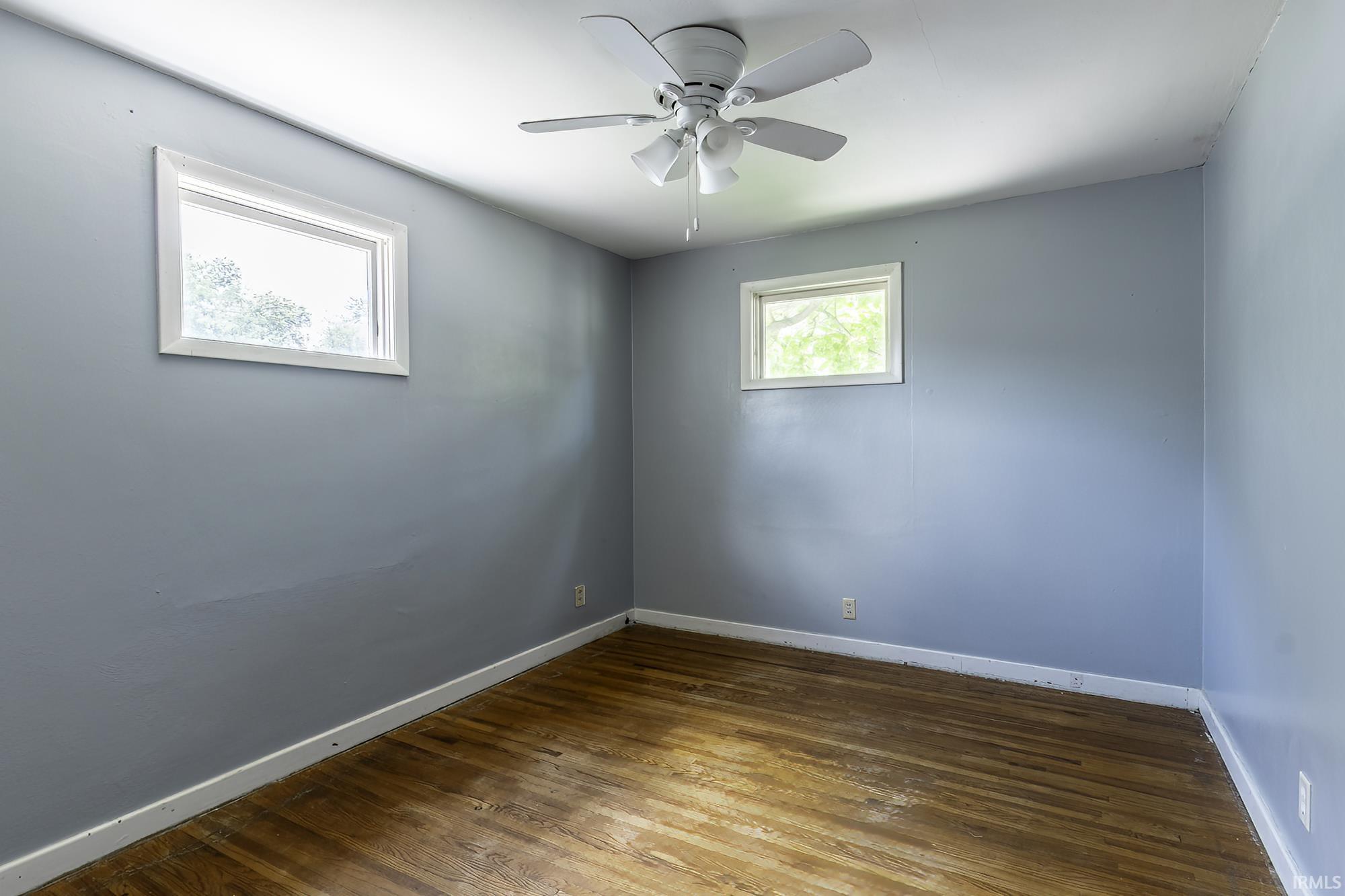 Room featuring wood-finish flooring, two windows with white trim, a ceiling fan with light fixture, and light blue wall paint