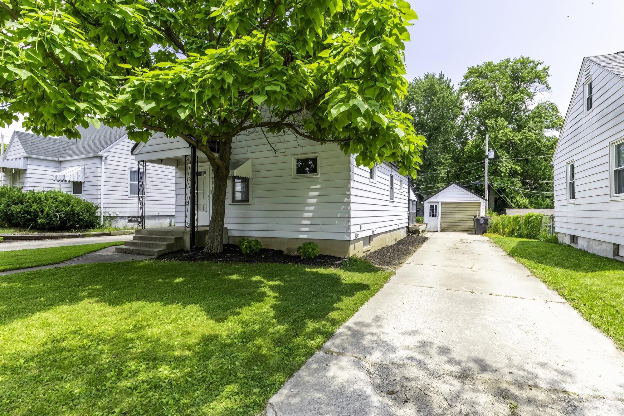 White siding residence featuring a concrete driveway, detached garage with wood-finish door, and mature front yard tree