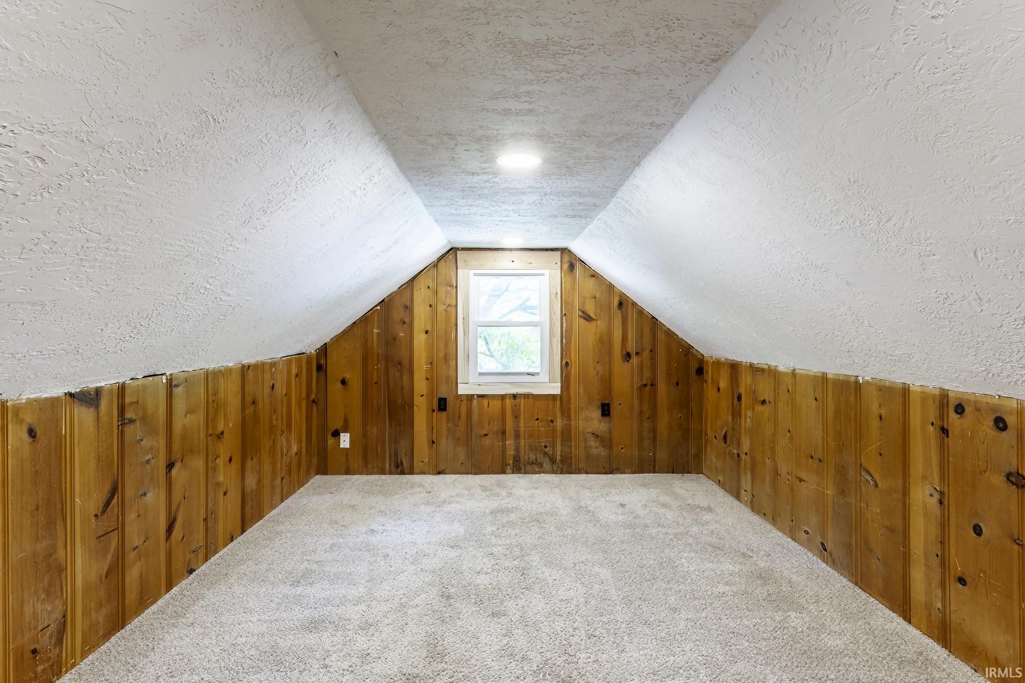 Attic space featuring light-toned carpeting, wood paneling, and a white framed window