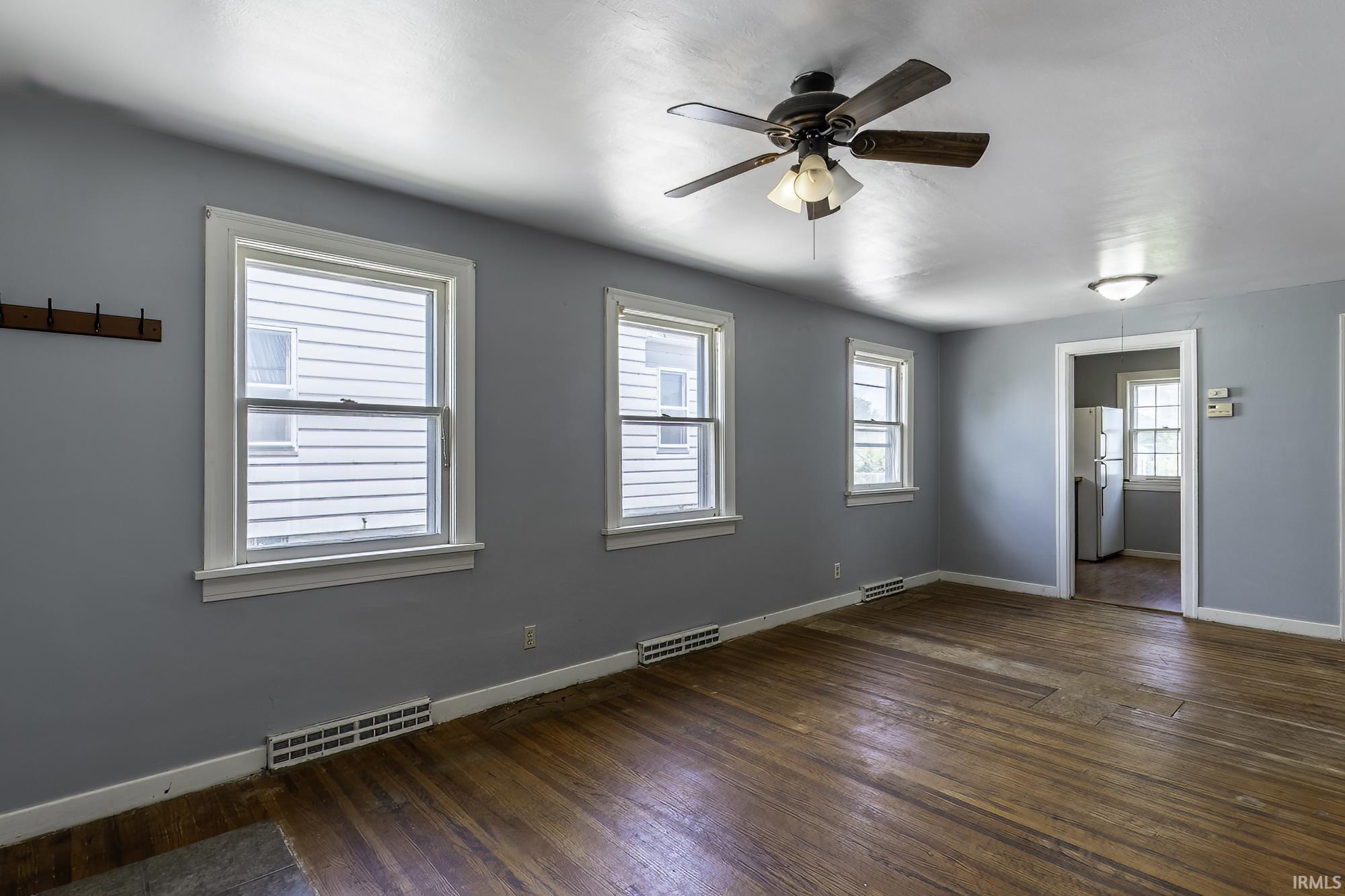 Spacious room featuring wood-finish flooring, three windows with white trim, a ceiling fan with light fixture, and a built-in coat rack