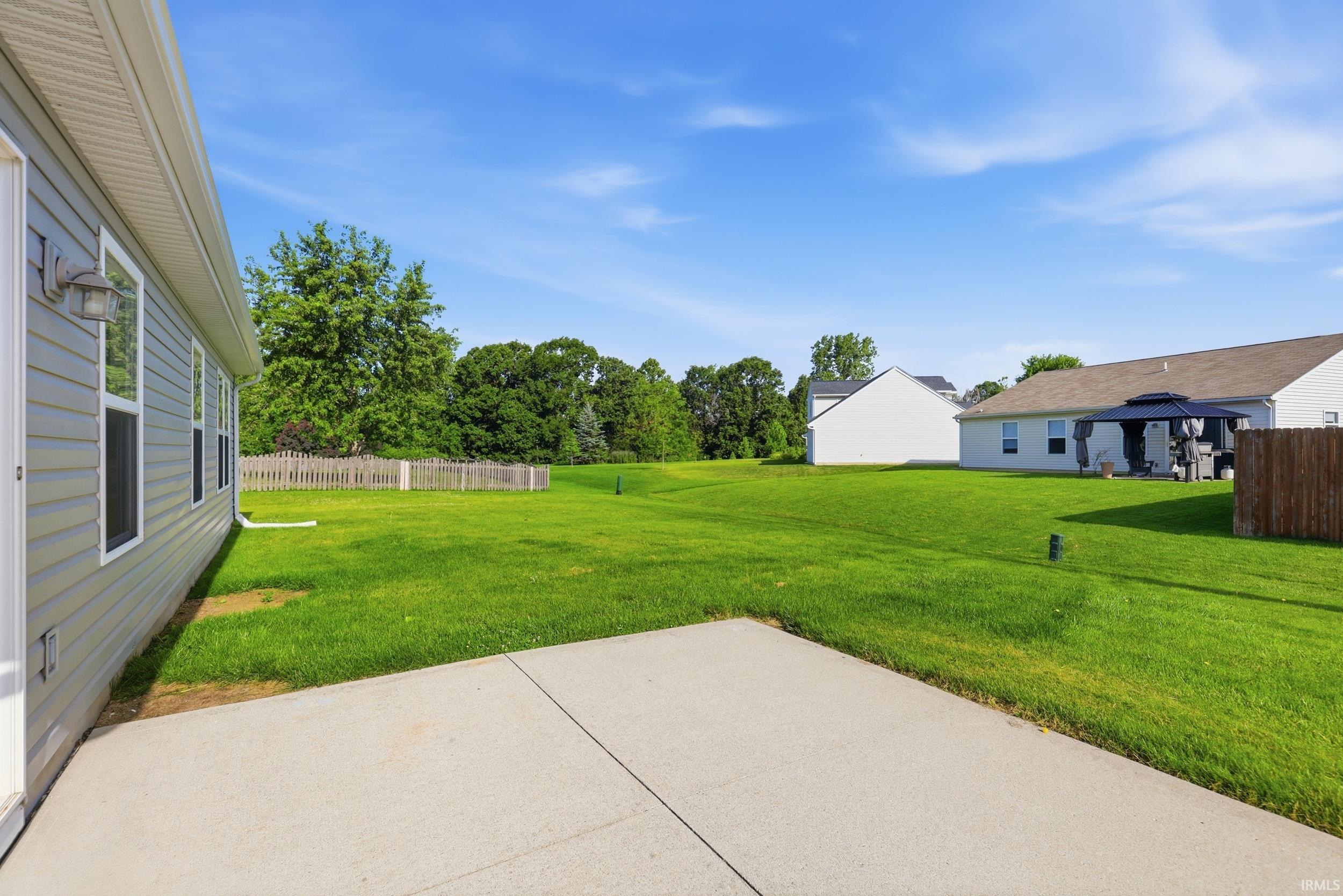Expansive green lawn with a concrete patio, featuring light gray horizontal siding, multiple windows, and a partial wooden fence
