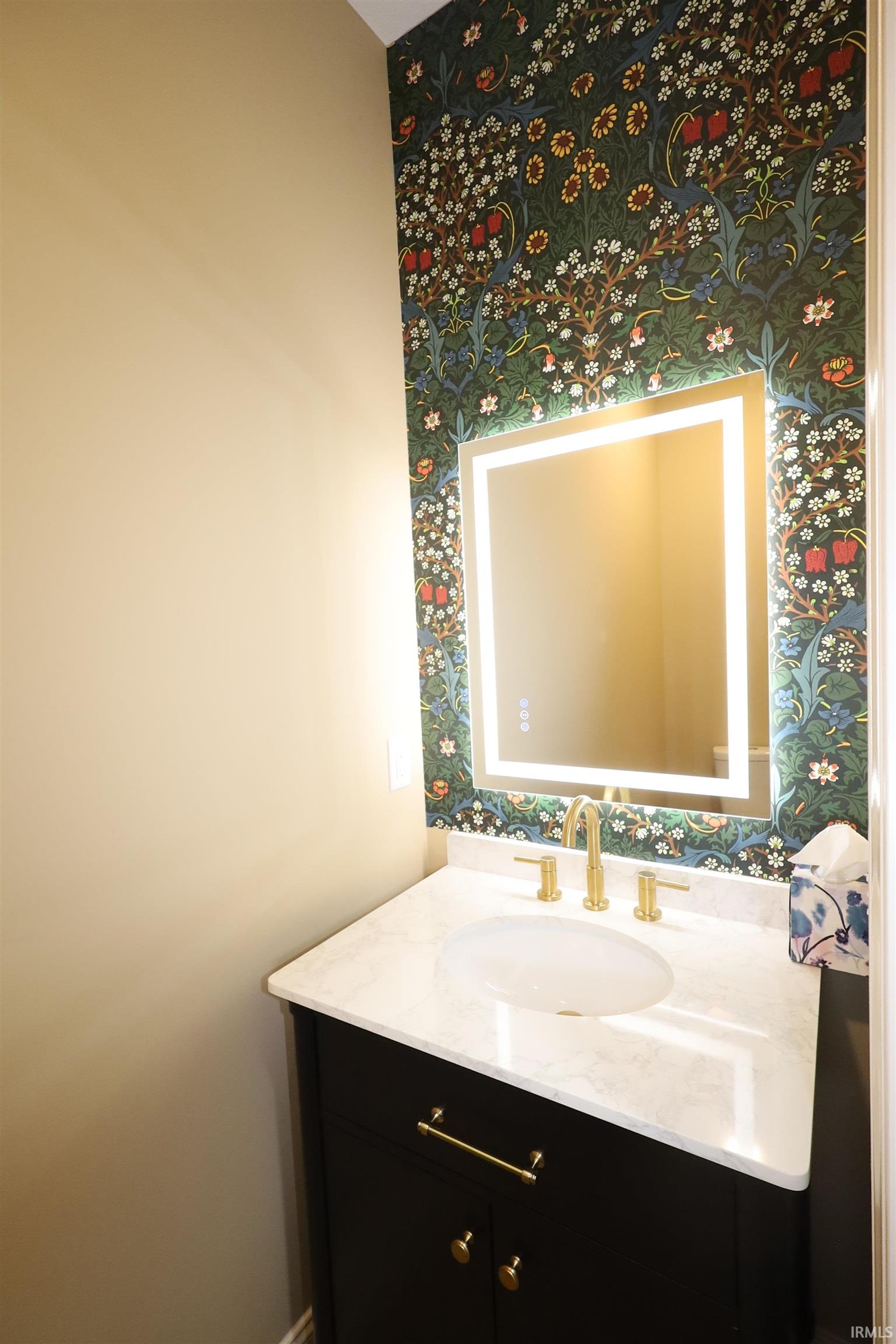 Powder room featuring a decorative botanical wallpaper accent wall, black vanity with gold hardware, integrated sink, and gold-finish faucet