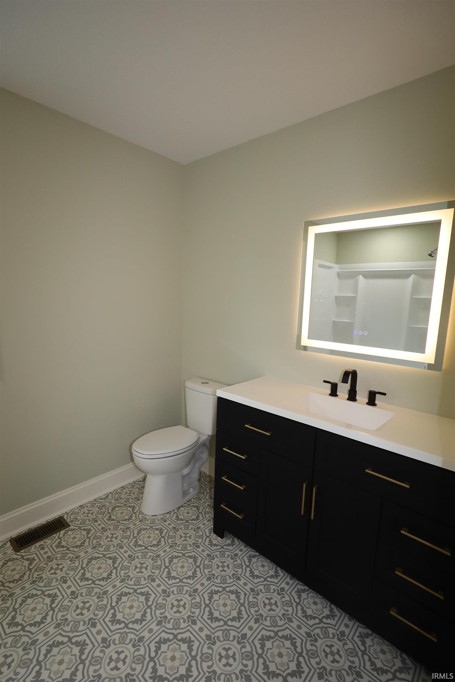 Bathroom featuring patterned floor tiling, a dark vanity with gold-tone hardware, a white countertop with an integrated sink, a back-lit mirror, and a white toilet