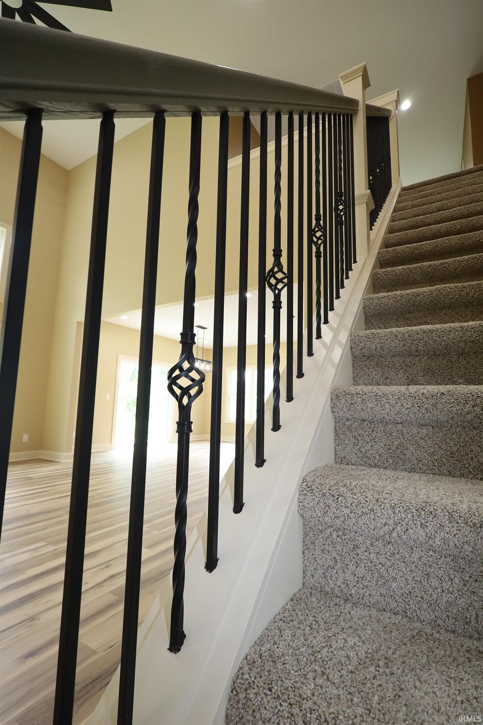 Staircase featuring wrought iron balusters with decorative elements, a dark-toned handrail, and light-toned carpeted treads