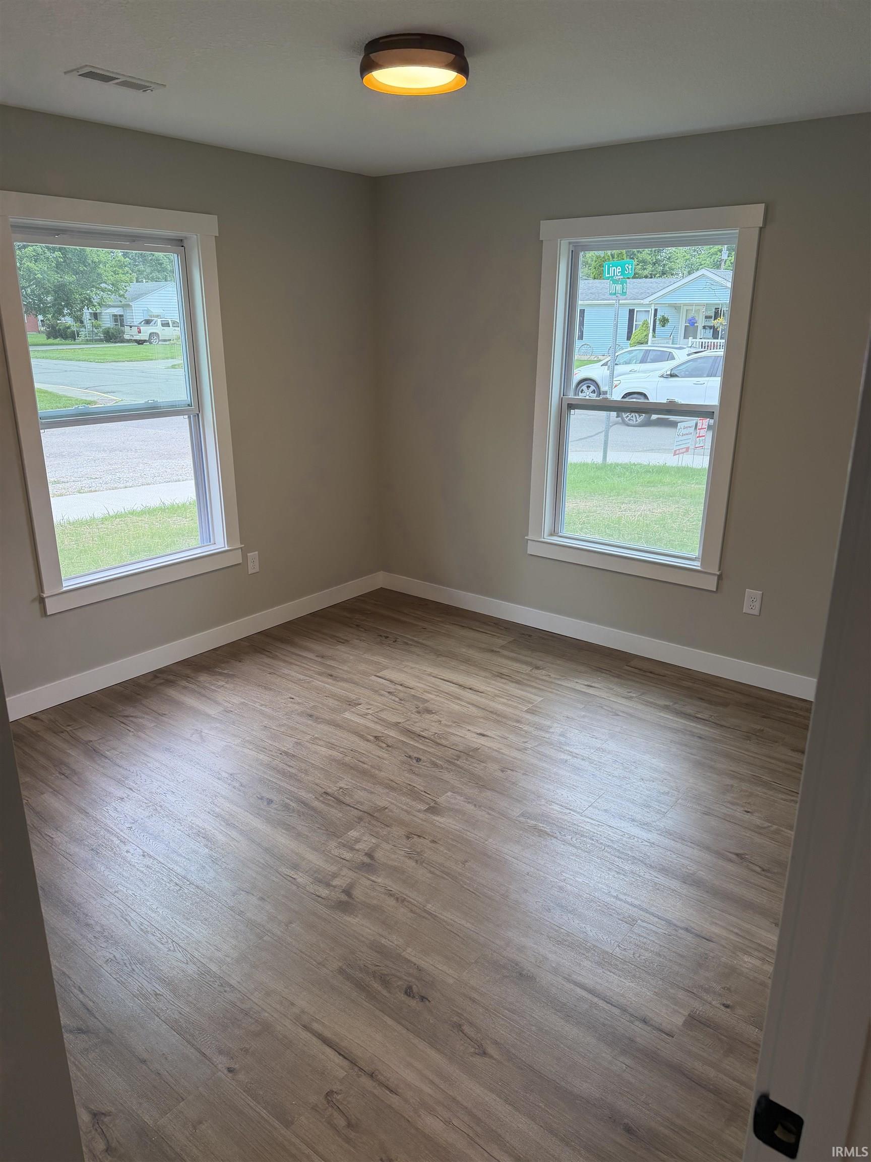 Spacious room featuring wood-finish flooring, two large windows with white trim, light gray walls, and a contemporary ceiling light fixture