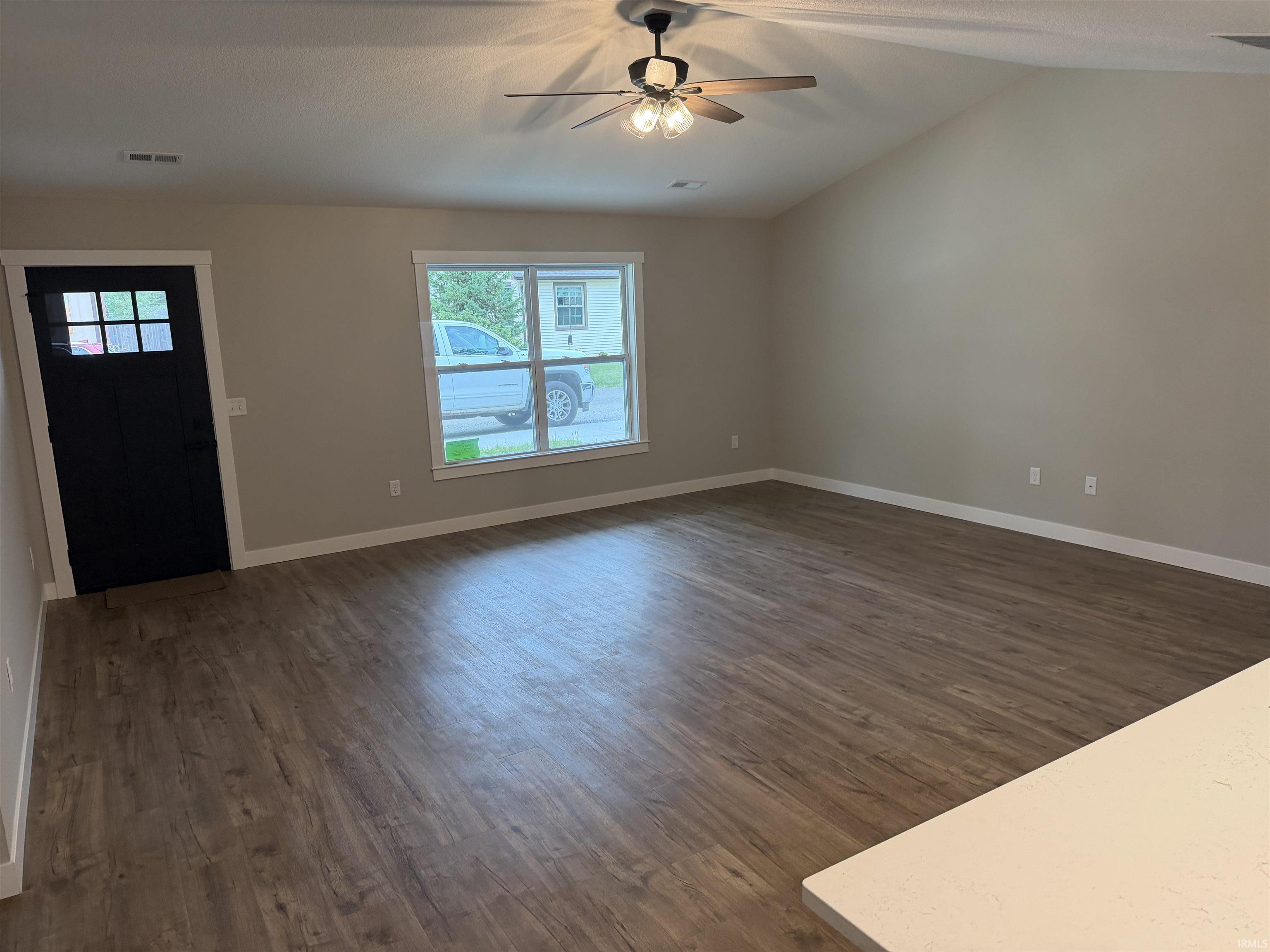 Spacious room featuring vaulted ceilings, wood-finish flooring, a contemporary ceiling fan with lighting, a multi-pane entry door, and a large window providing natural illumination