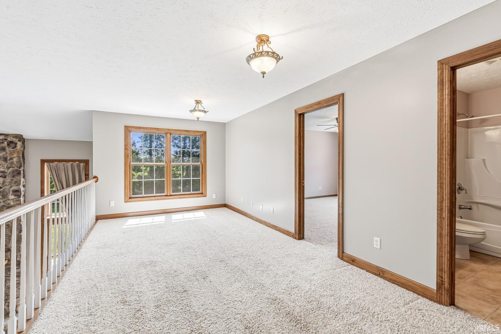 Carpeted loft area featuring a double window with wood trim, two ceiling-mounted light fixtures, a stone architectural element, and a white baluster railing