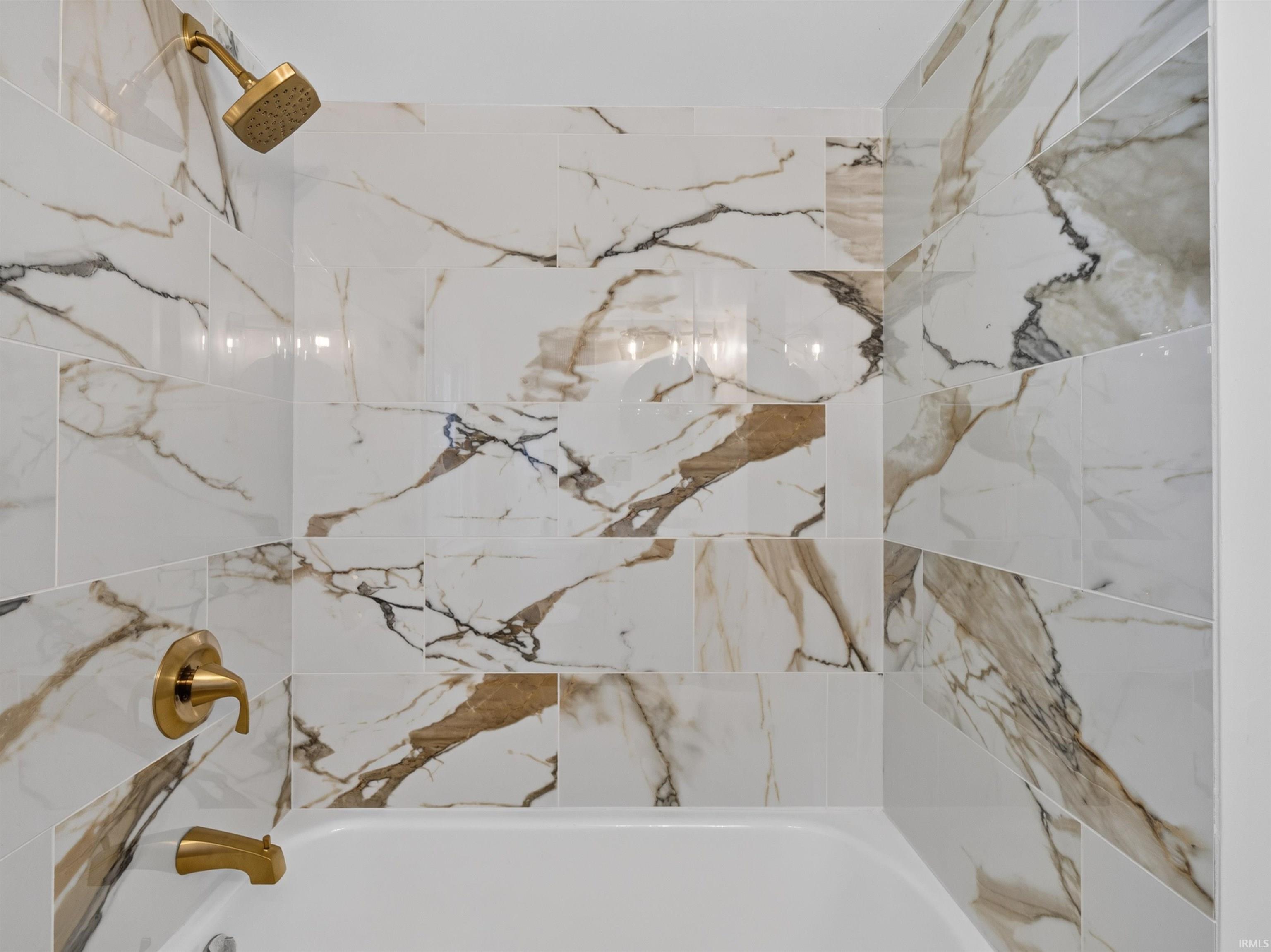 Bathtub and shower combination featuring large format marble-look wall tiles, a gold-finish showerhead, and matching gold-finish tub filler and handle
