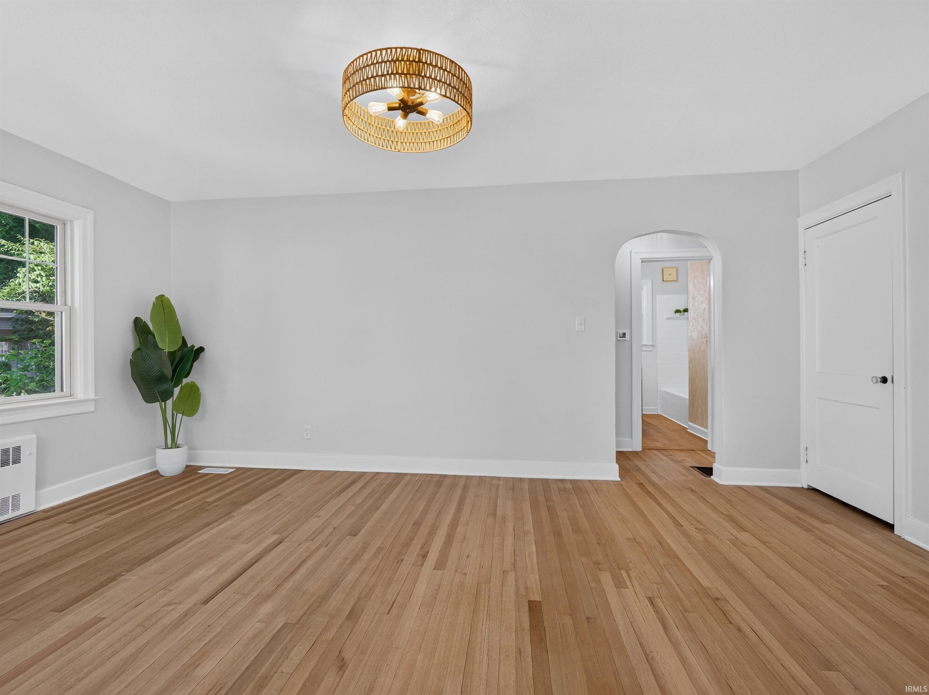 Hardwood flooring throughout a room featuring a modern woven ceiling light fixture, white trim, a single-pane window, and an arched doorway