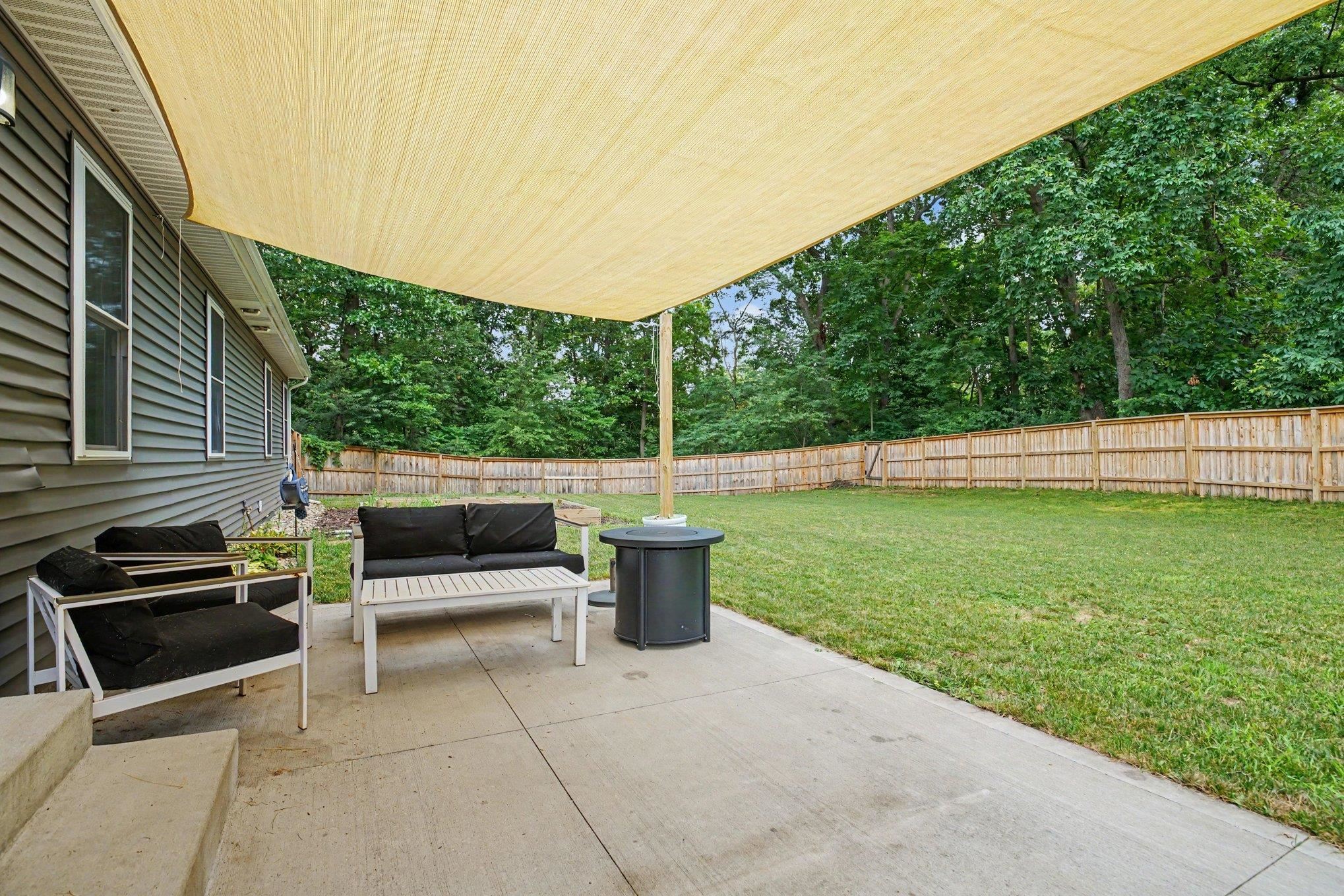 Expansive concrete patio offering a large shade sail, siding exterior, mature tree line, and wood privacy fencing