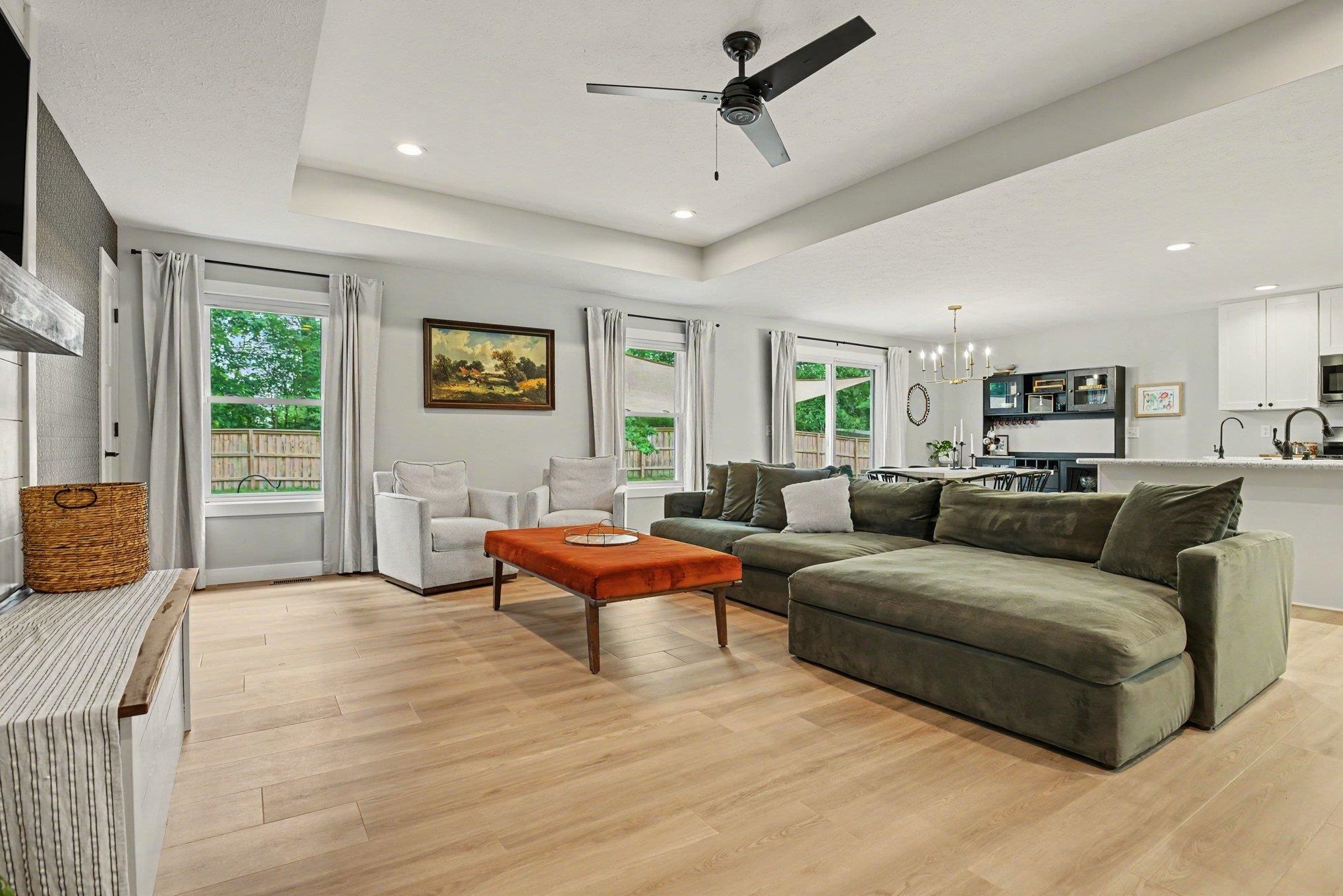 Open-concept living area featuring light wood-finish flooring, a tray ceiling with a ceiling fan, recessed lighting, and multiple large windows
