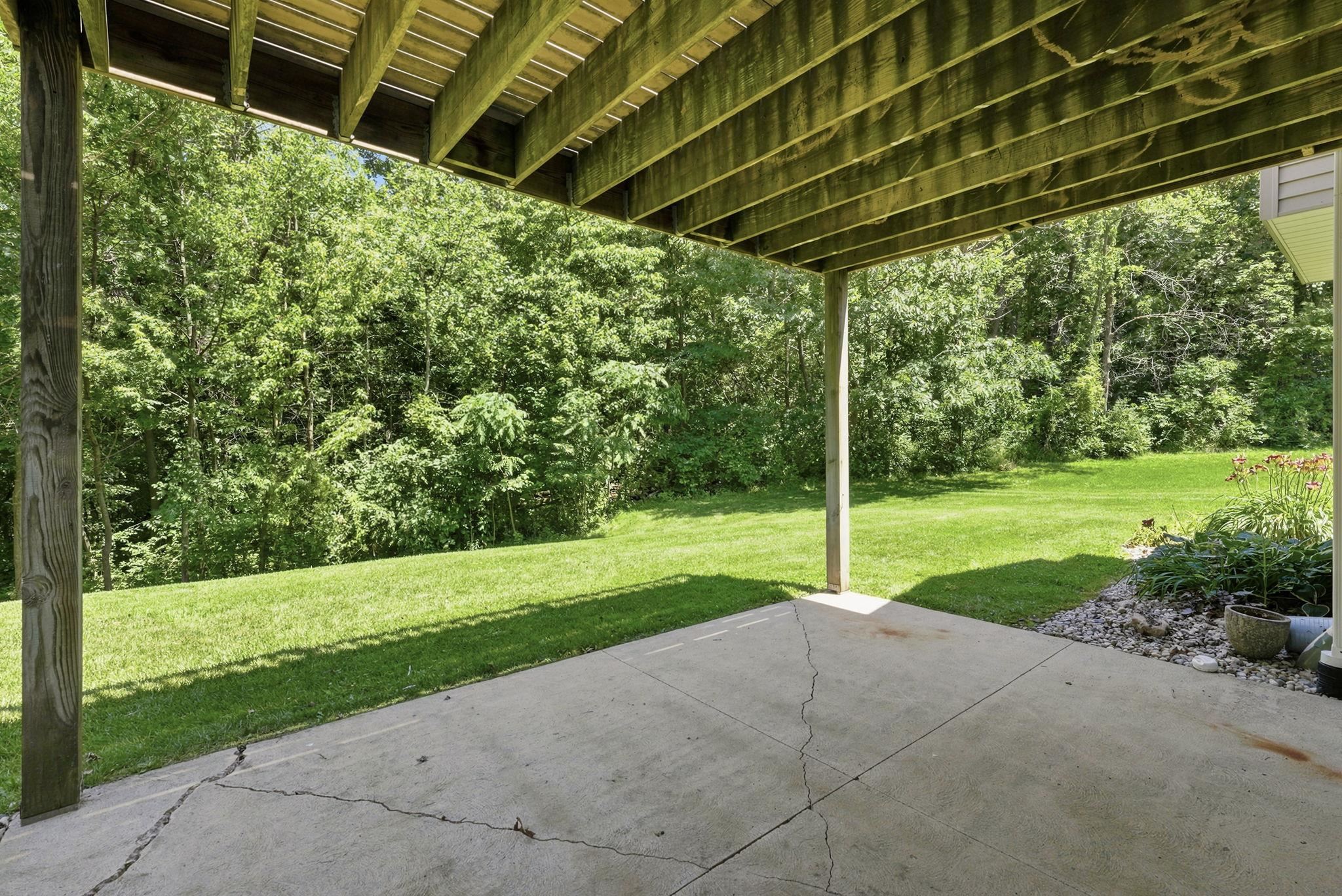 Covered patio with concrete slab and exposed wood joists overhead, bordered by a manicured lawn and mature trees