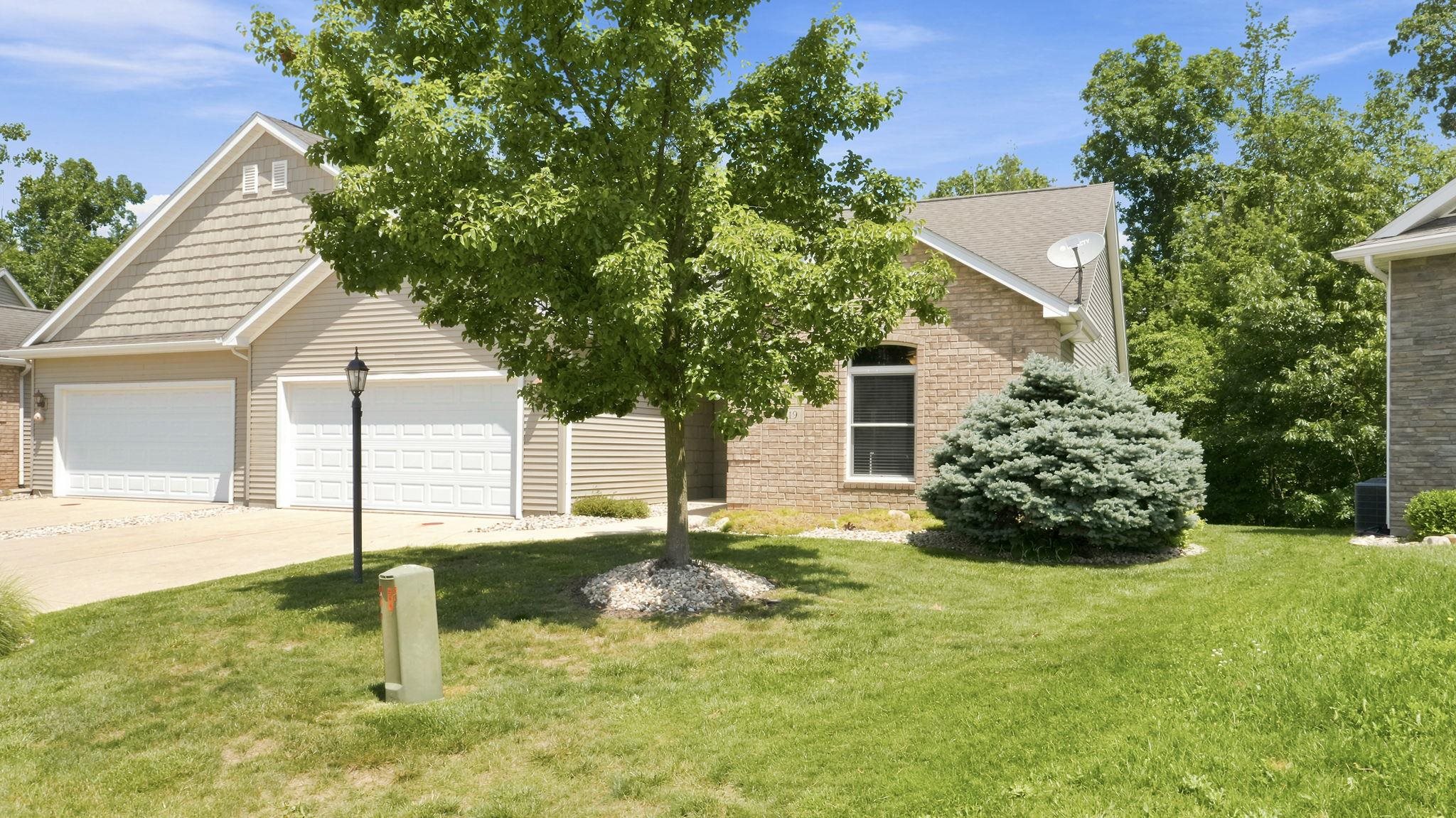 Exterior featuring a combination of brick and tan horizontal siding, a two-car garage with white paneled doors, a paved driveway, and mature landscaping