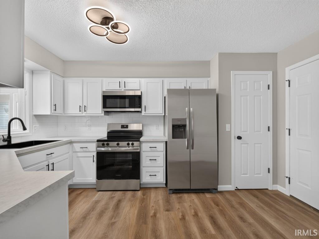 Kitchen featuring white cabinetry, stainless steel appliances, a black sink, wood-finish flooring, and a unique contemporary ceiling light fixture