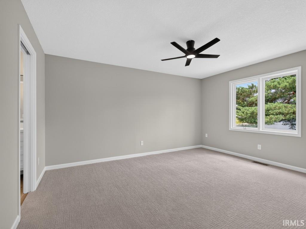 Spacious room featuring neutral carpeting, light gray walls, white trim, a modern black ceiling fan with integrated lighting, and a dual-pane window offering a verdant outdoor landscape