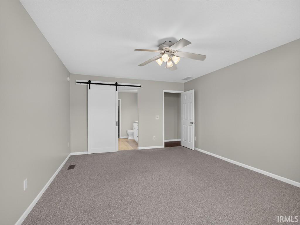 Spacious room featuring neutral paint, gray carpeting, a ceiling fan with integrated lighting, white baseboards, and a modern barn door entry to an additional space