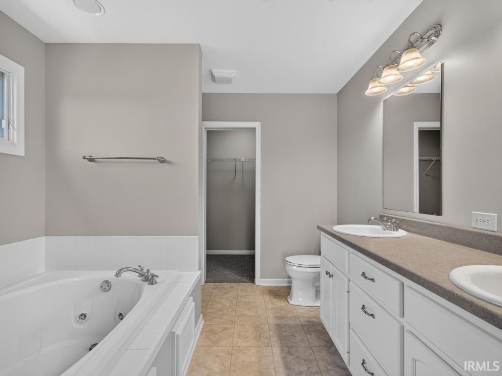Bathroom featuring a jetted soaking tub with tile surround, a double vanity with integrated sinks, a toilet, and a walk-in closet with wire shelving