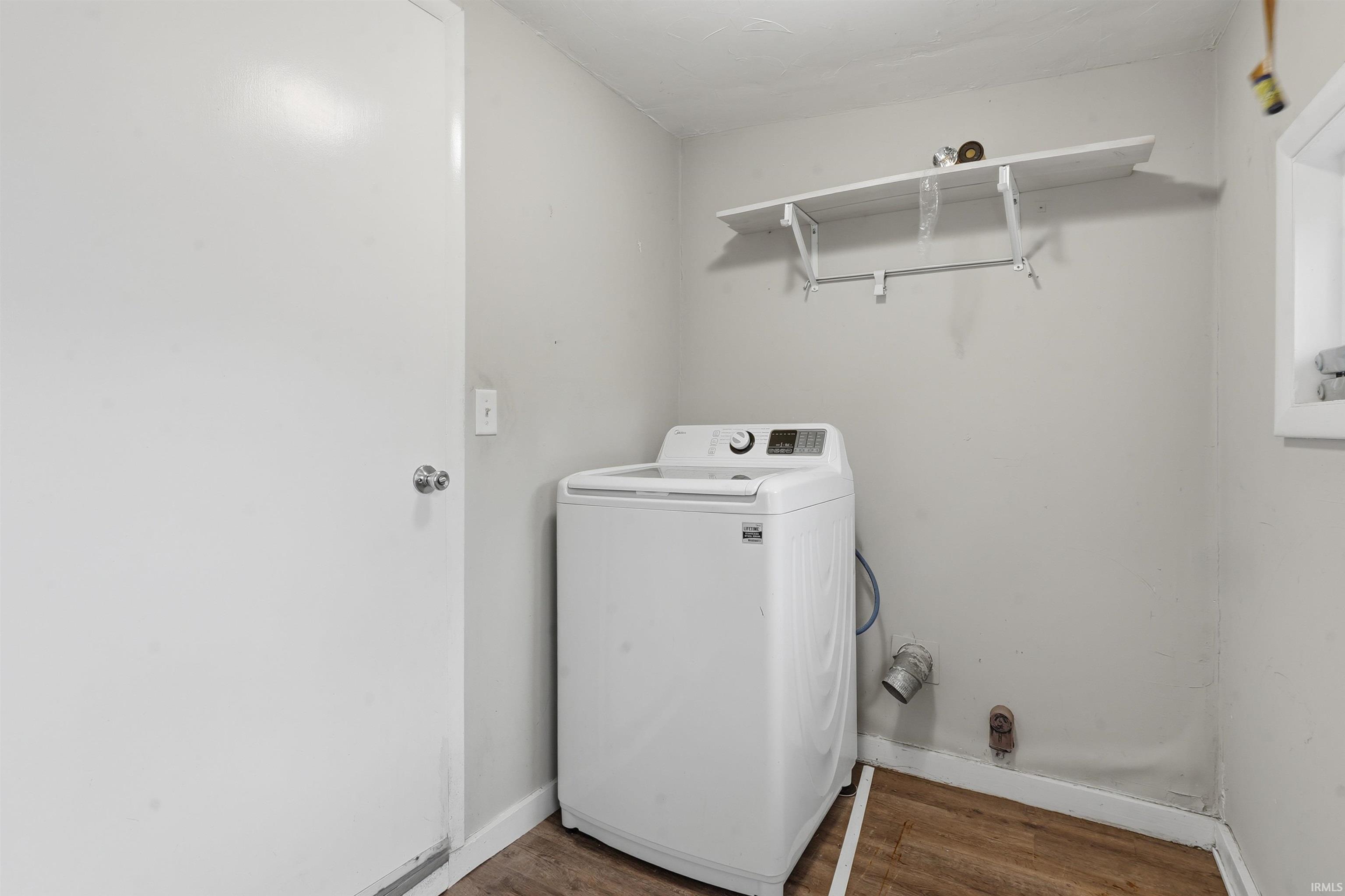 Dedicated laundry area featuring wood-finish flooring, a white interior door with a silver knob, a wall-mounted white shelf, and a white-framed window