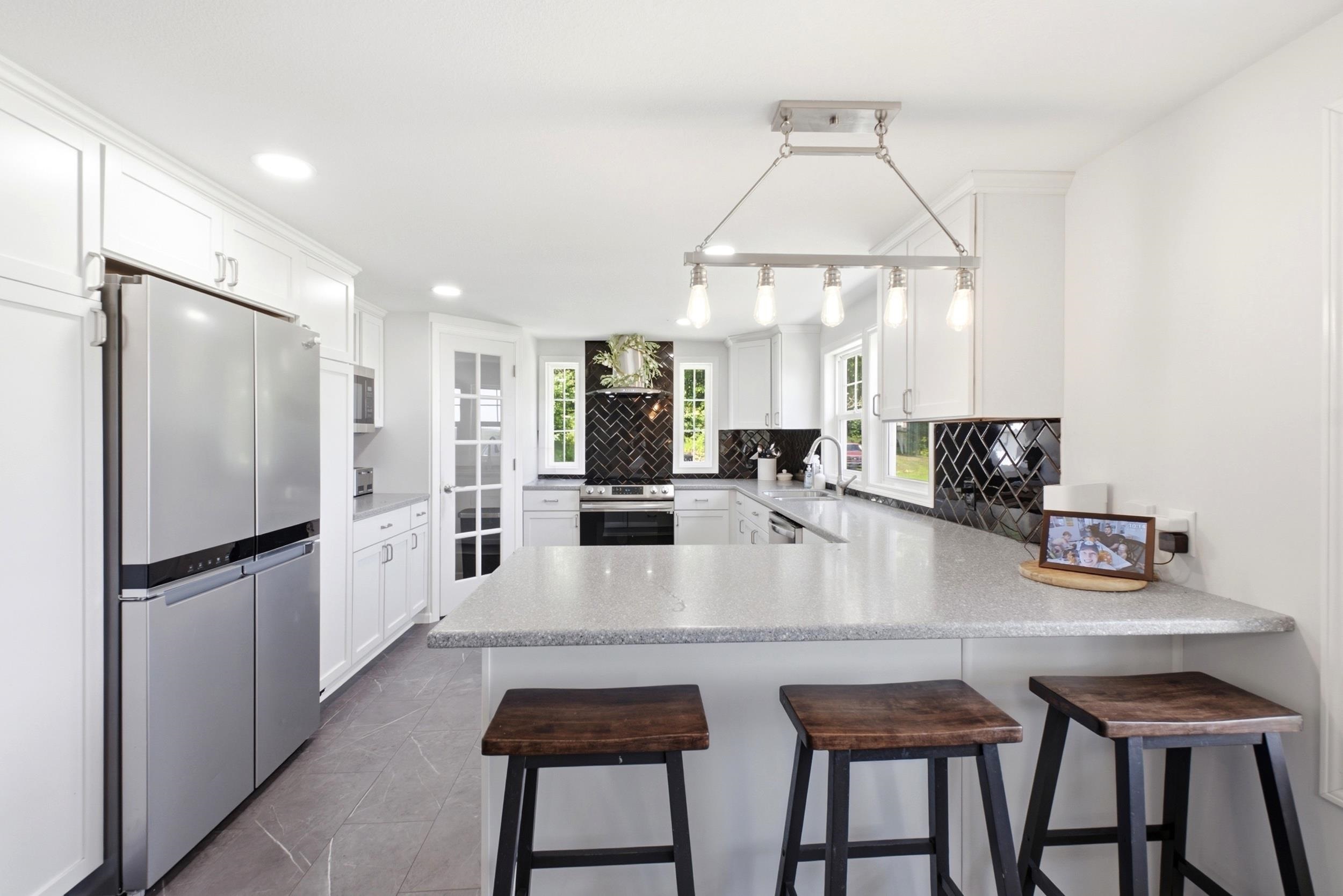 Modern kitchen featuring a spacious island with a light-toned countertop, sleek stainless steel appliances, white shaker cabinetry, a herringbone pattern dark tile backsplash, and recessed lighting throughout