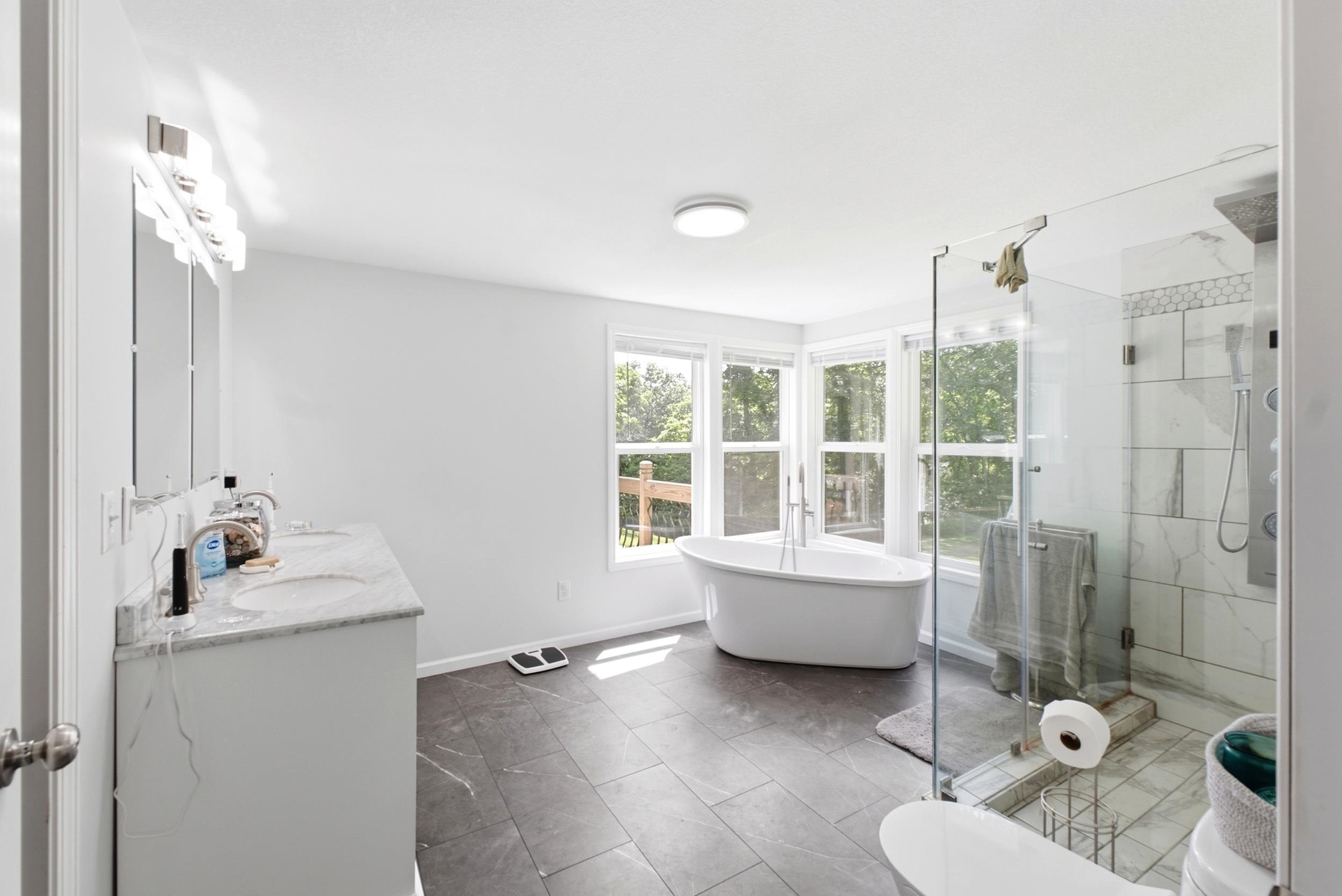 Spacious bathroom featuring a freestanding soaking tub, walk-in shower with clear glass enclosure, dual vanity with undermount sinks and stone countertop, and dark-toned floor tiling