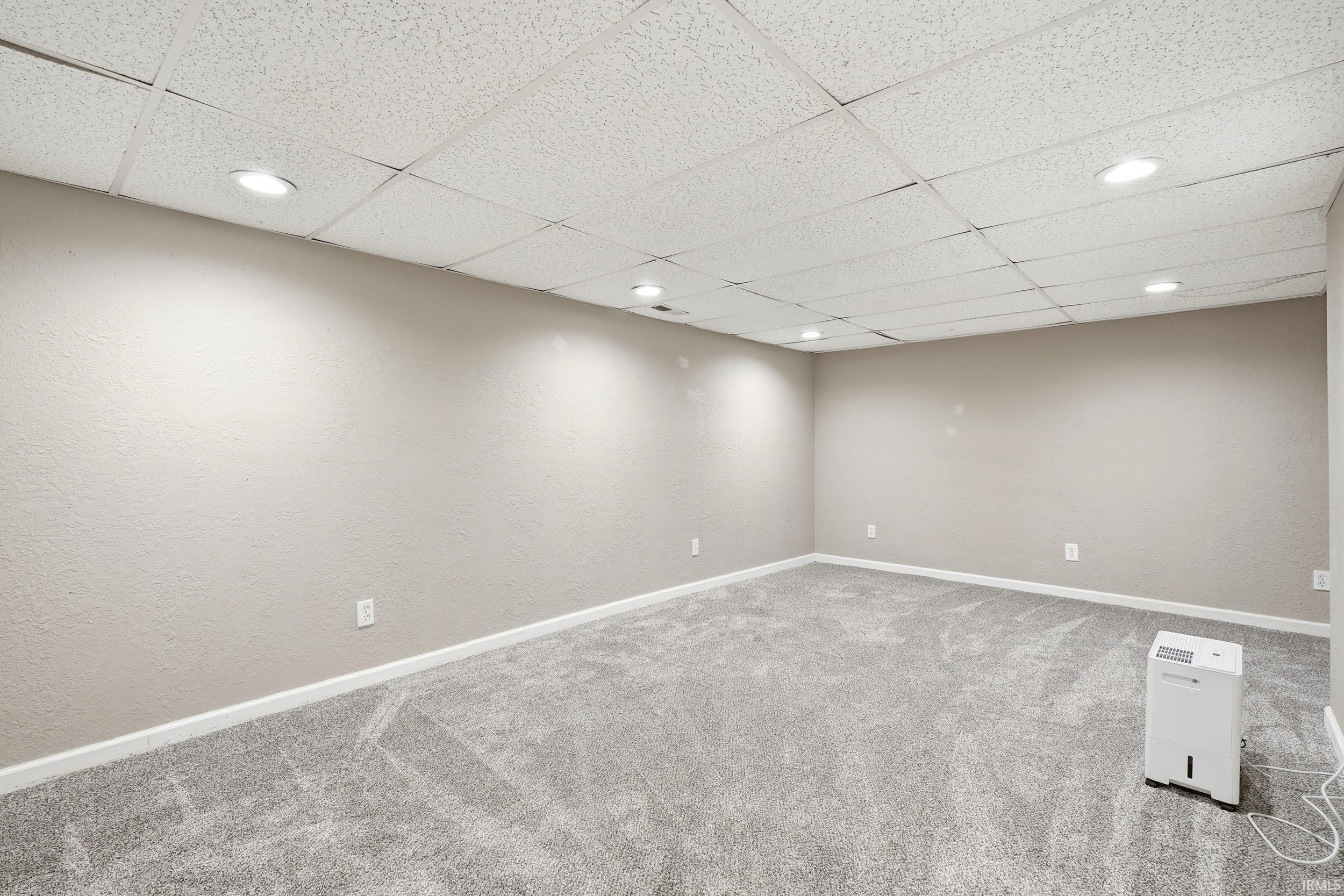 Spacious room featuring neutral painted walls, light grey carpet, white baseboards, and a drop ceiling with recessed lighting