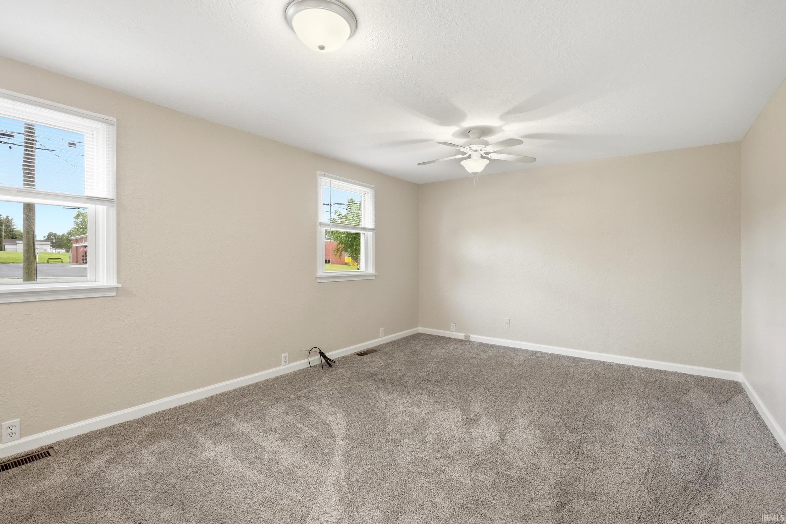 Spacious room featuring neutral paint, gray carpeting, a ceiling fan with integrated lighting, and dual windows allowing natural illumination