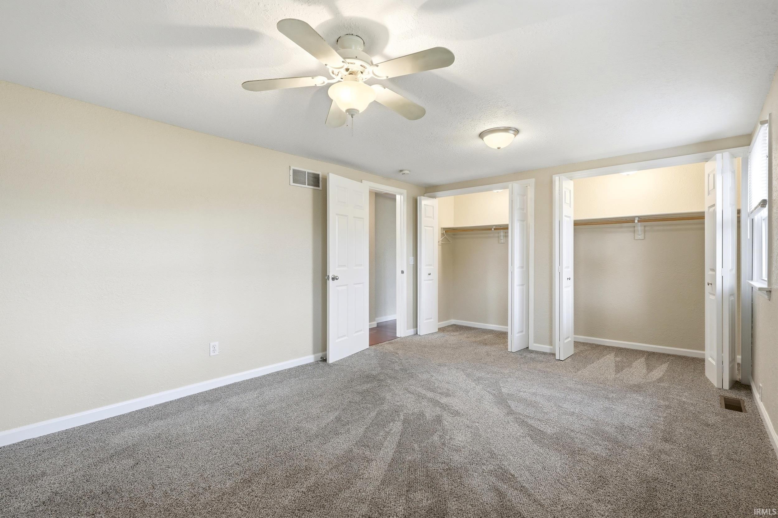 Spacious room featuring neutral carpeting, two bi-fold door closets, a ceiling fan with integrated lighting, and additional flush-mount ceiling lighting