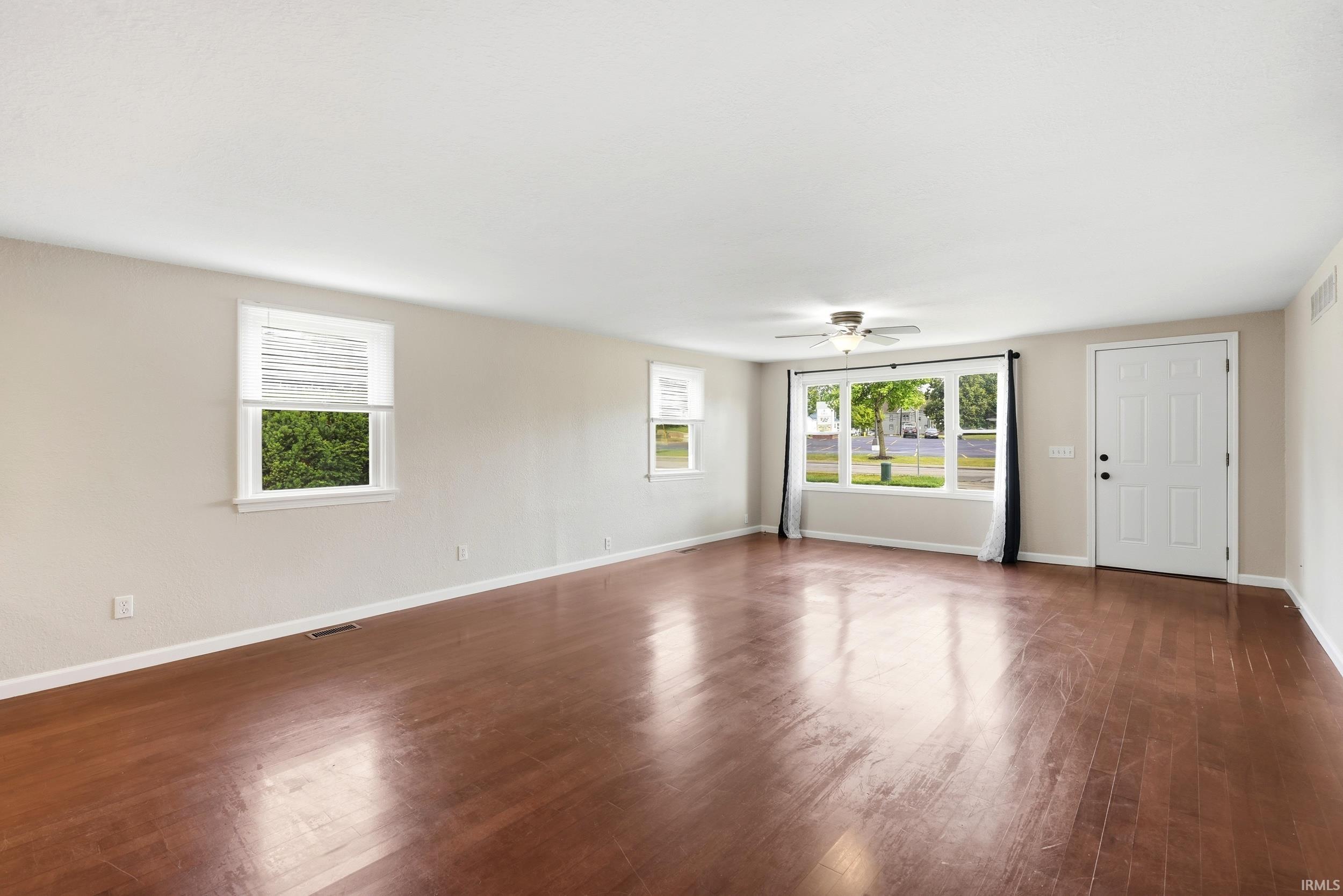Spacious room featuring wood-finish flooring, light-toned walls, multiple windows with blinds, a front entry door, and a ceiling fan