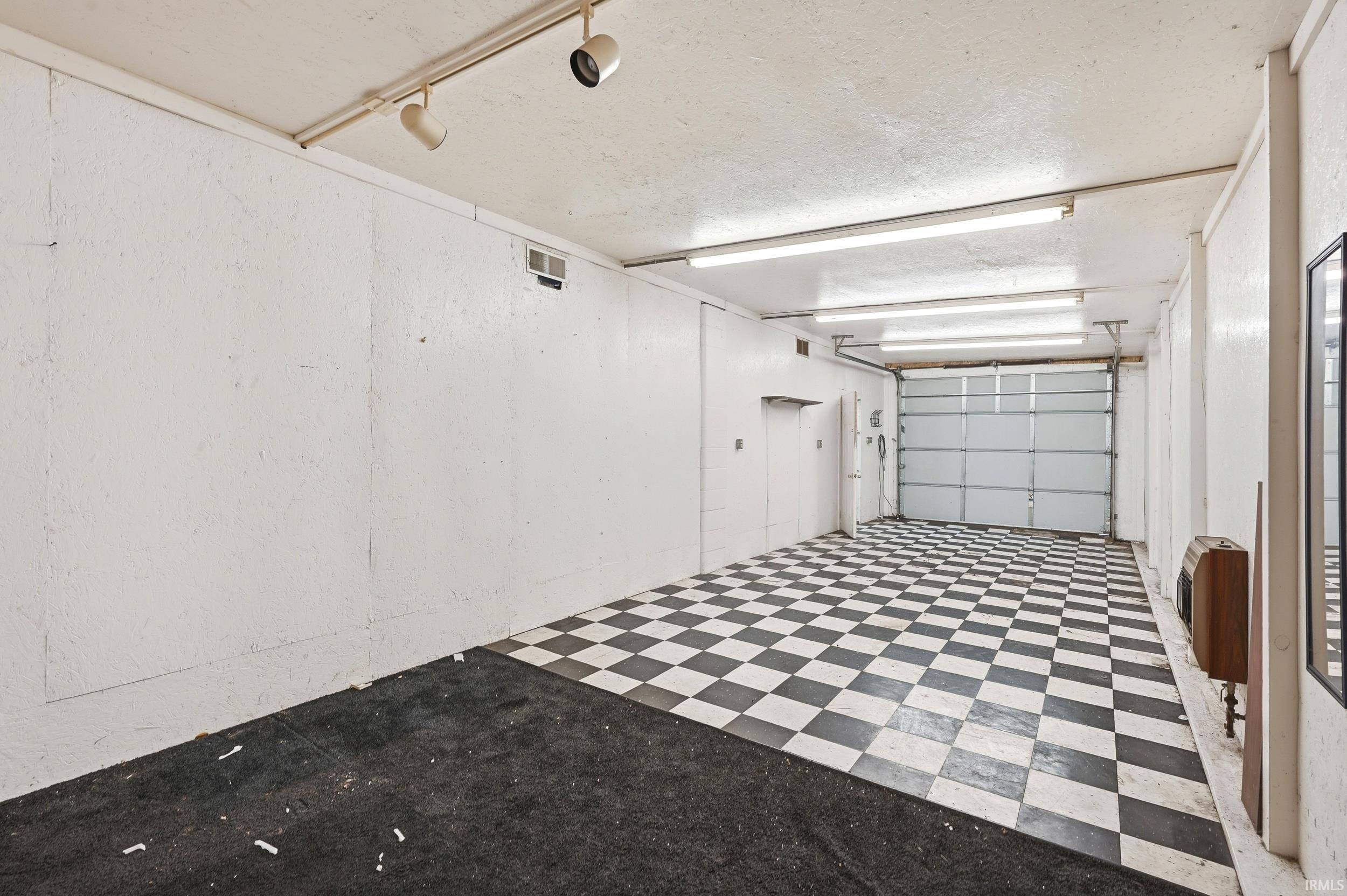 Spacious garage featuring a checkerboard floor, overhead fluorescent lighting, track lighting, and a roll-up garage door