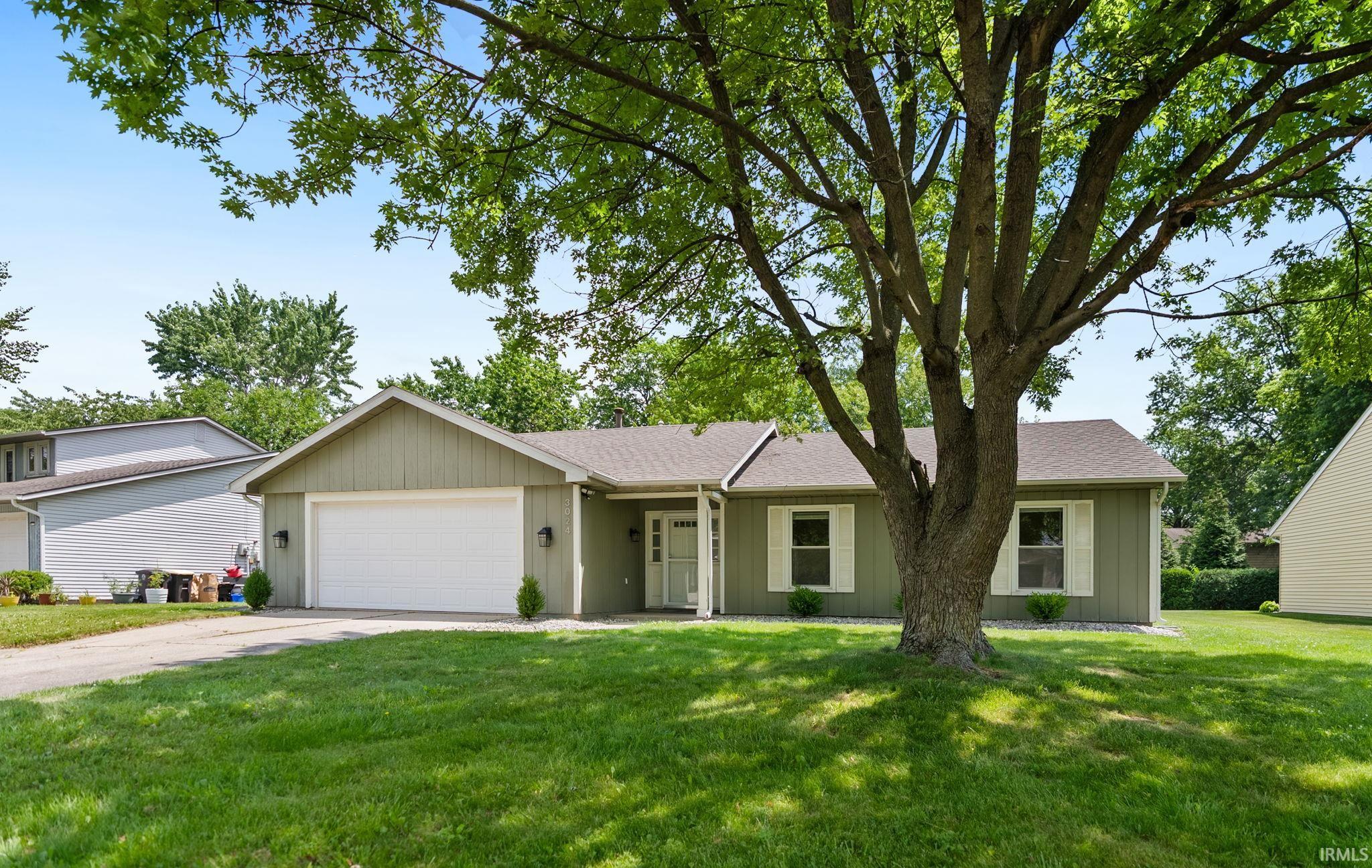 Ranch-style residence featuring a prominent front lawn, mature shade tree, attached two-car garage, and a welcoming front entrance