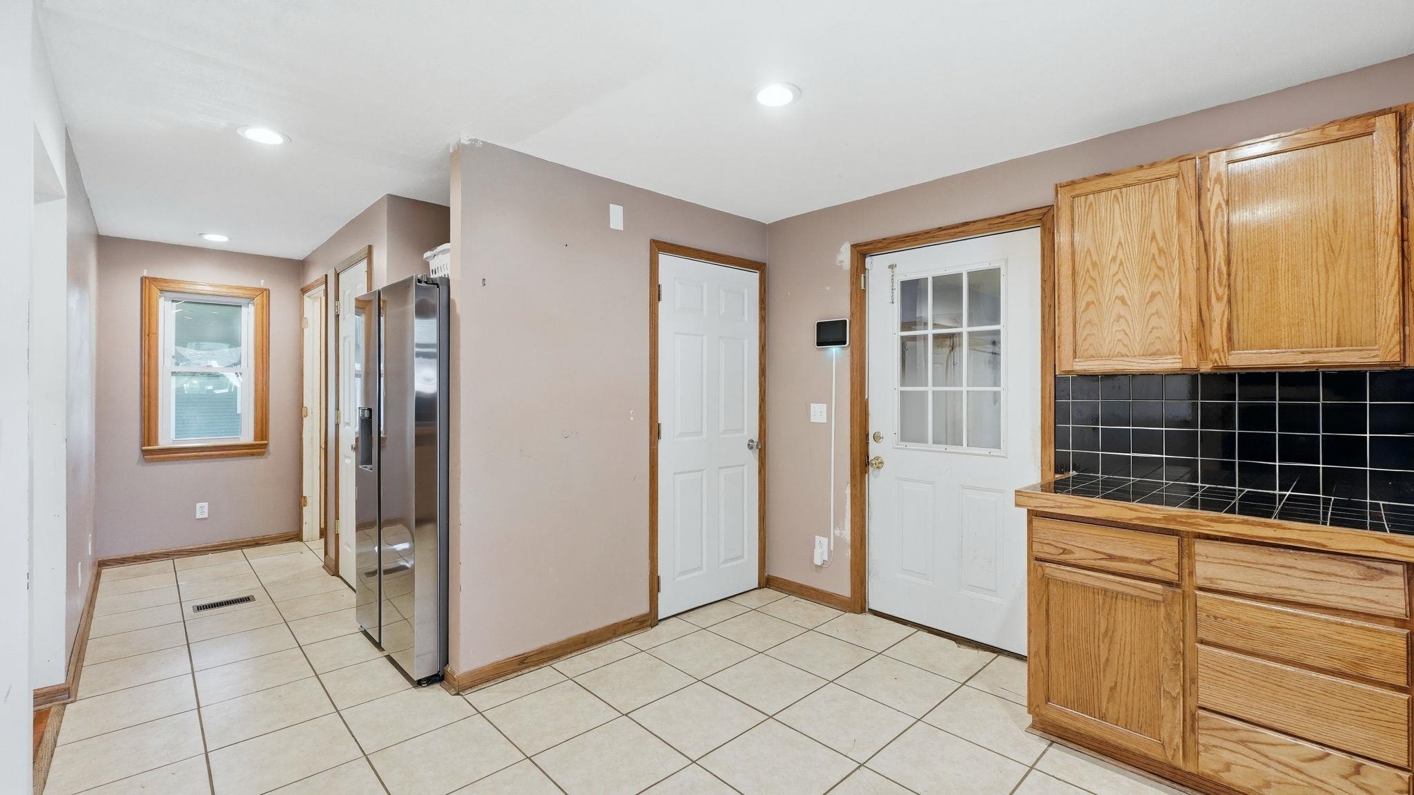 Kitchen area featuring light-toned tile flooring, wood-finish cabinetry, black tile backsplash, and a built-in stainless steel refrigerator