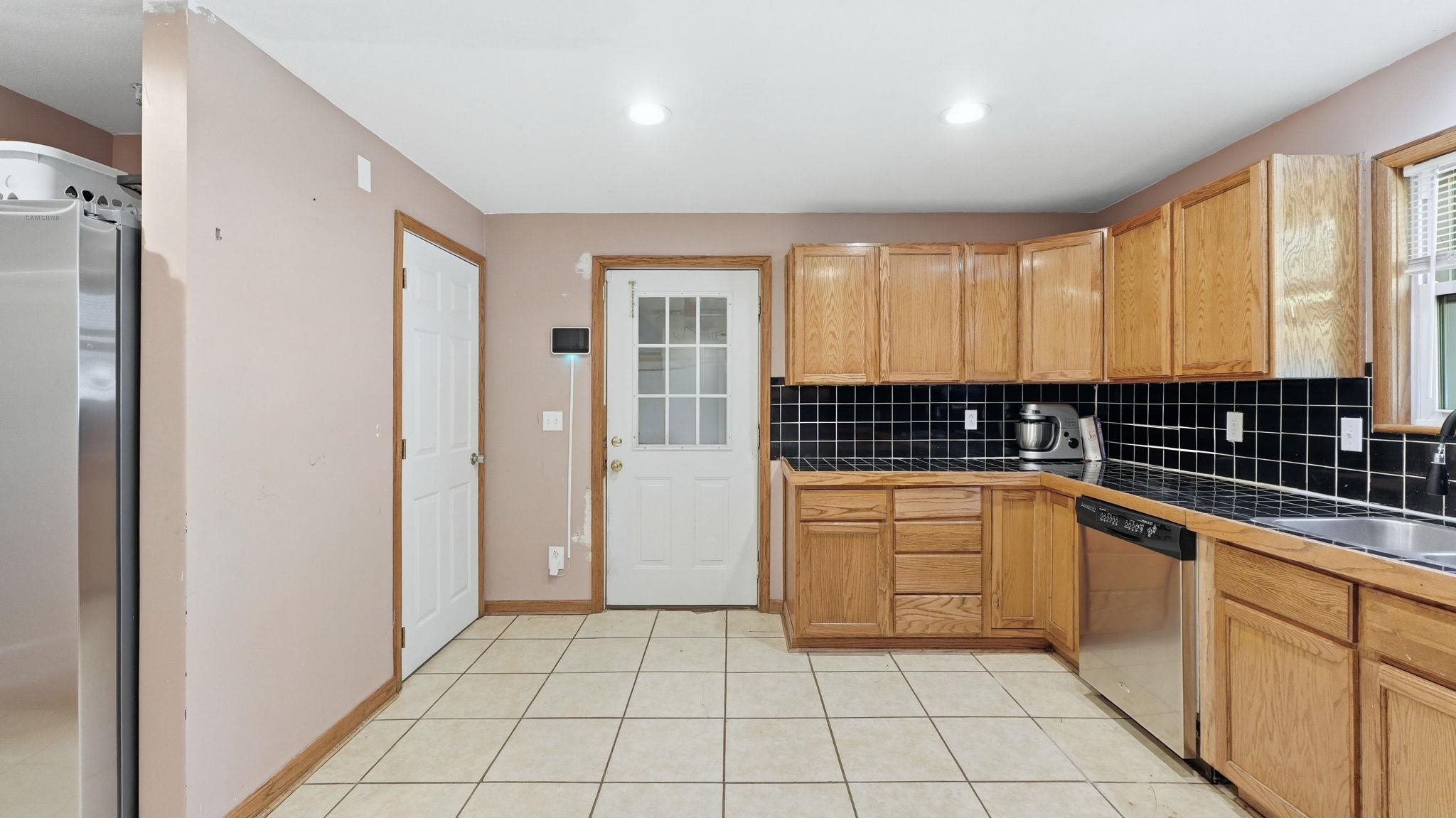 Kitchen featuring light wood cabinetry, black subway tile backsplash, stainless steel dishwasher, and tile flooring